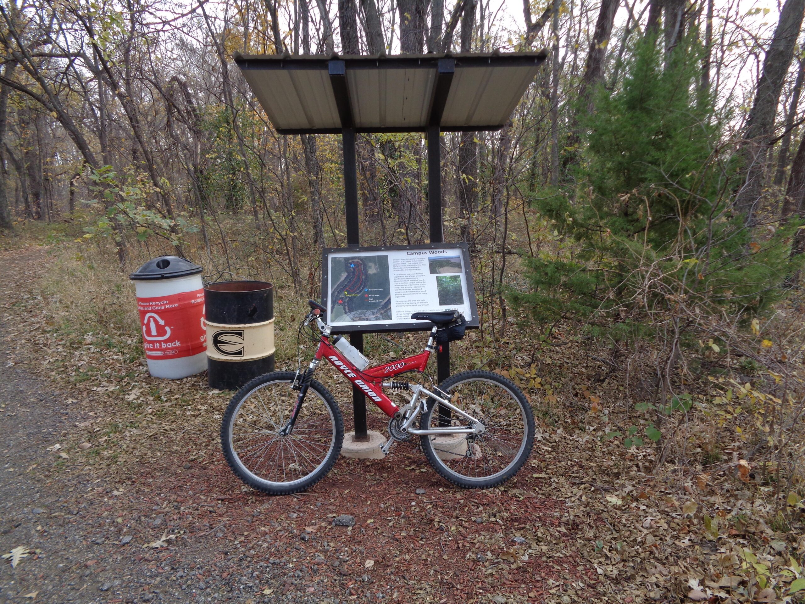 A mountain bike parked next to a trail sign in a wooded area, with a recycling bin nearby. The scene features autumn foliage with trees in the background, indicating a nature trail or park setting. ESU Trail mountain bike trail.