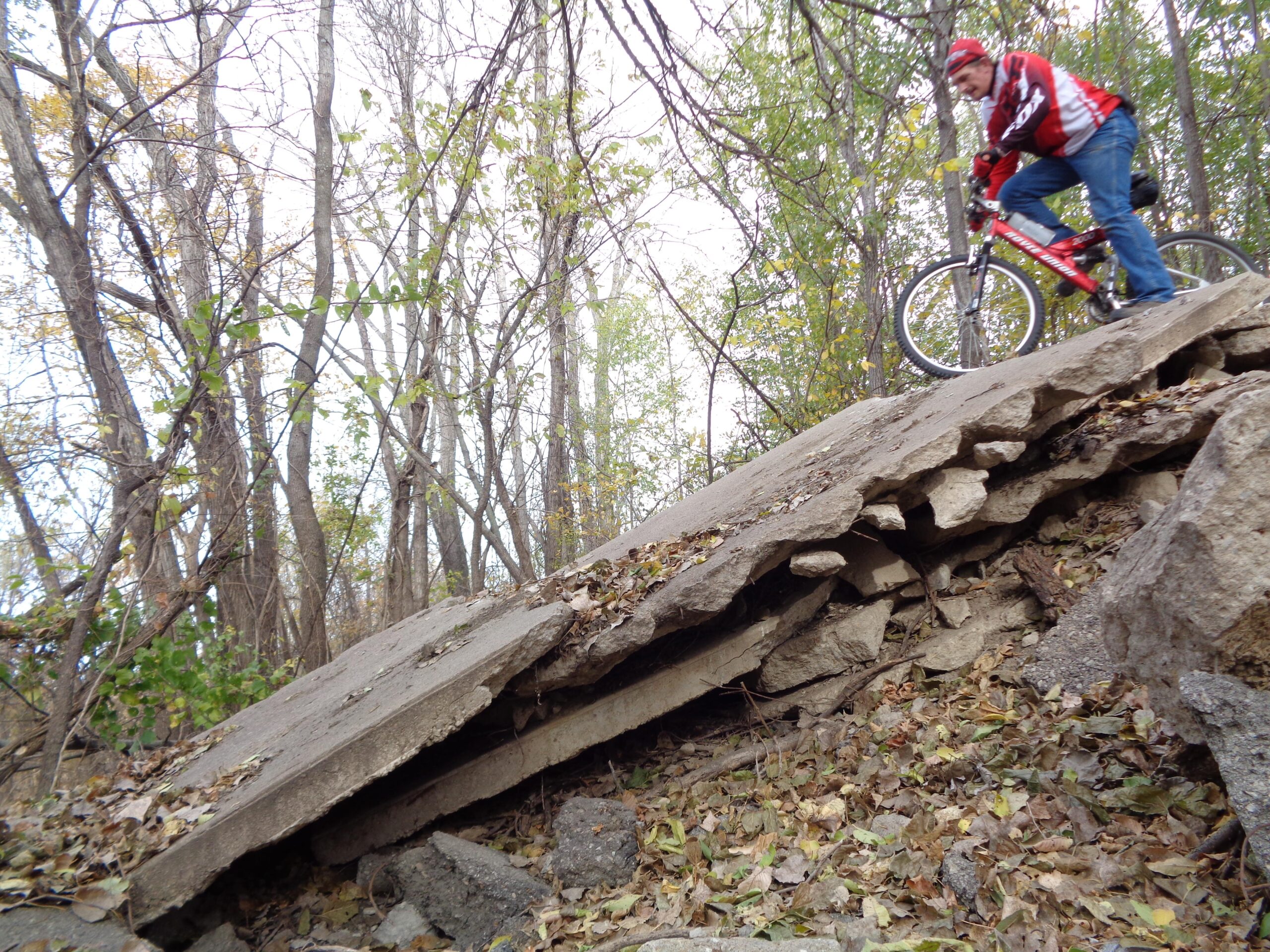 A cyclist navigating down a steep, rocky slope covered in autumn leaves, surrounded by bare trees. The cyclist is wearing a red and white jersey and is focused on controlling the bike as it descends the uneven terrain. ESU Trail mountain bike trail.