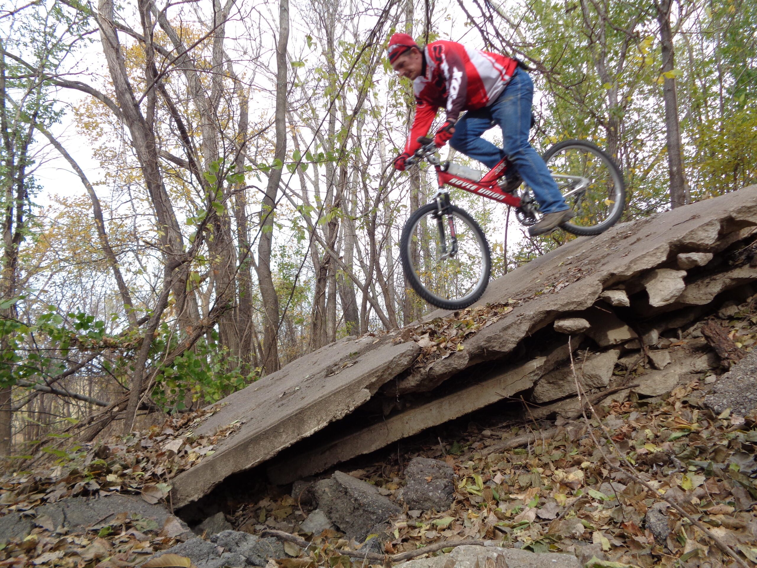 A cyclist expertly navigates a rocky descent, riding a red mountain bike over uneven terrain surrounded by trees and autumn foliage. ESU Trail mountain bike trail.