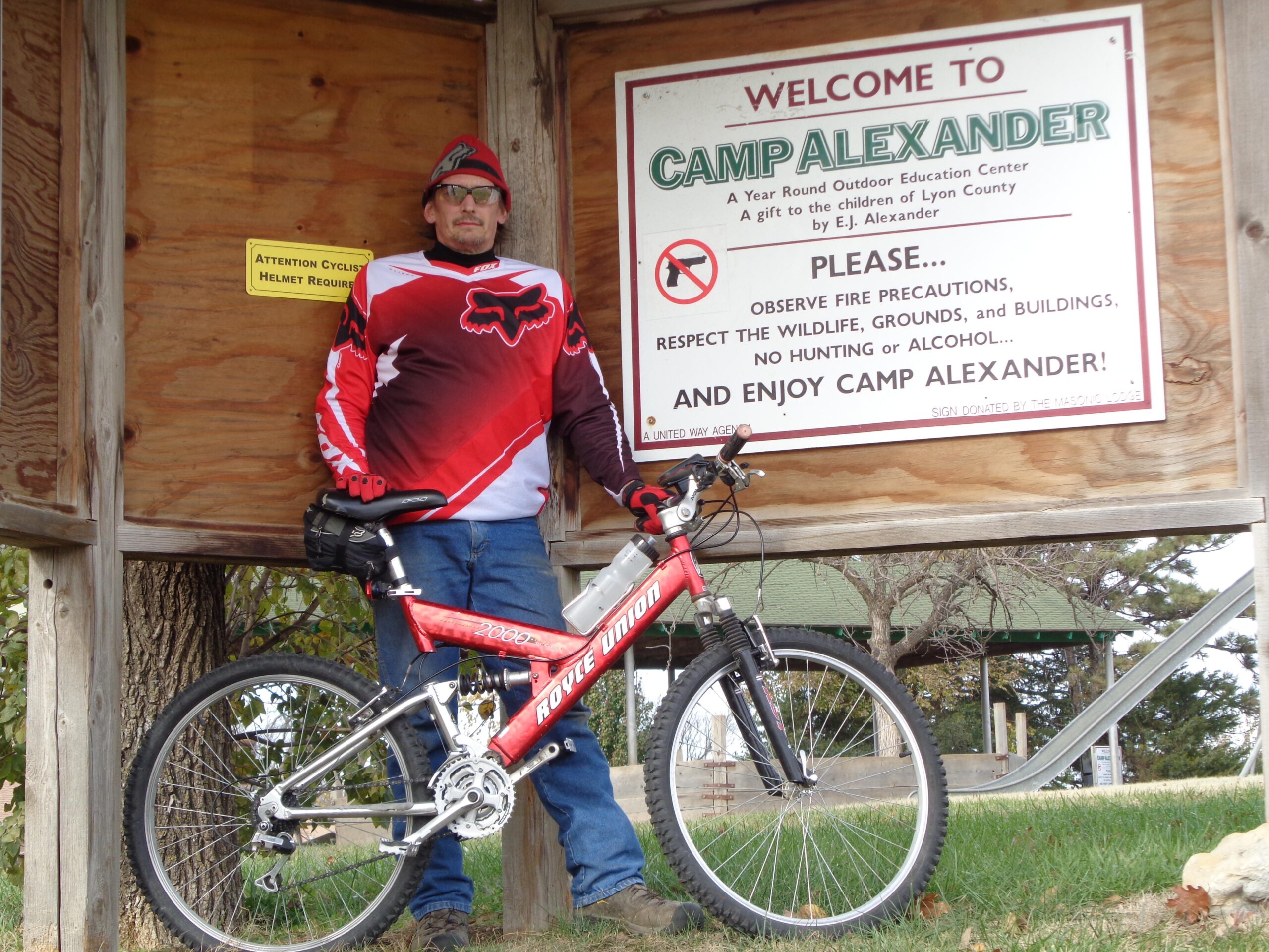A person in a red and black cycling jersey and a beanie stands next to a wooden sign that reads "Welcome to Camp Alexander," which outlines safety and behavior rules for the outdoor education center. The individual is holding a mountain bike, with the bike resting against the wooden structure. The background features trees and grassy areas typical of a camp setting. Camp Alexander Trails mountain bike trail.