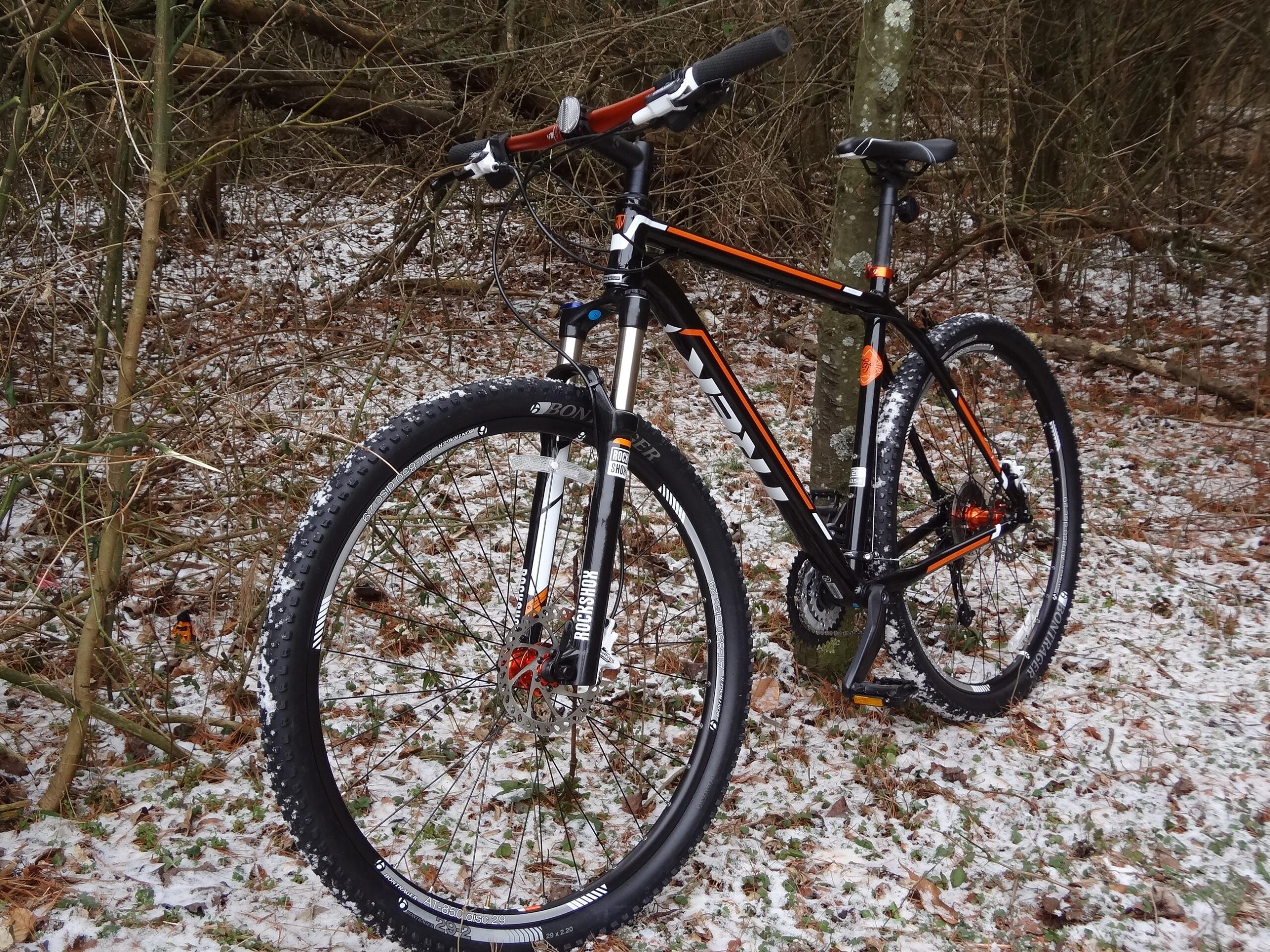 Trek Mamba: Mountain bike leaning against a tree in a forest setting, with a dusting of snow on the ground and bike tires. The bike features a black and orange frame with thick tires designed for rugged terrain.