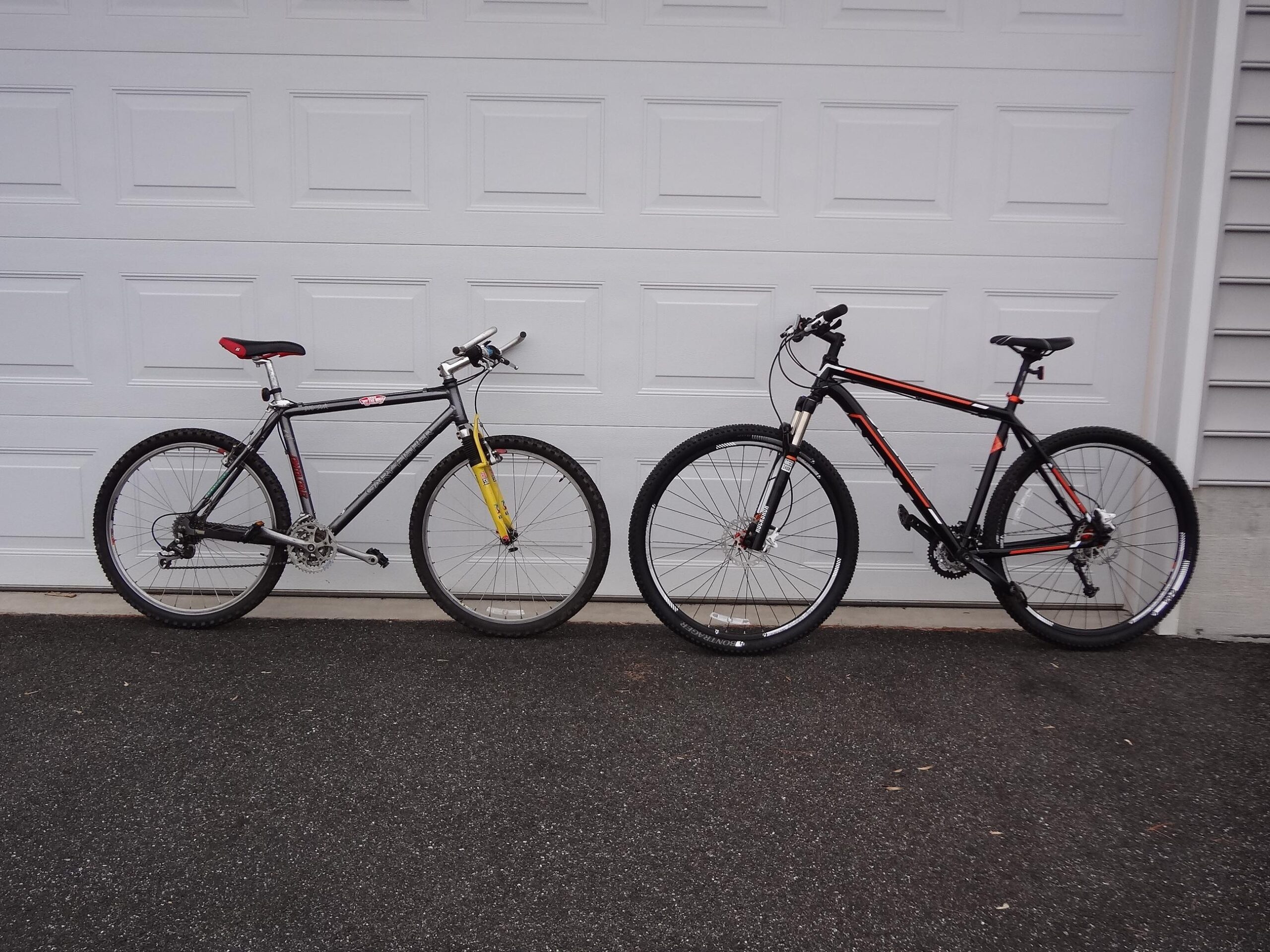 Gary Fisher Big Sur: Two mountain bikes parked side by side in front of a closed garage door. The bike on the left features a black frame with yellow fork and red accents, while the bike on the right has a black frame with red and white details. The ground is paved and the background includes a plain garage door.