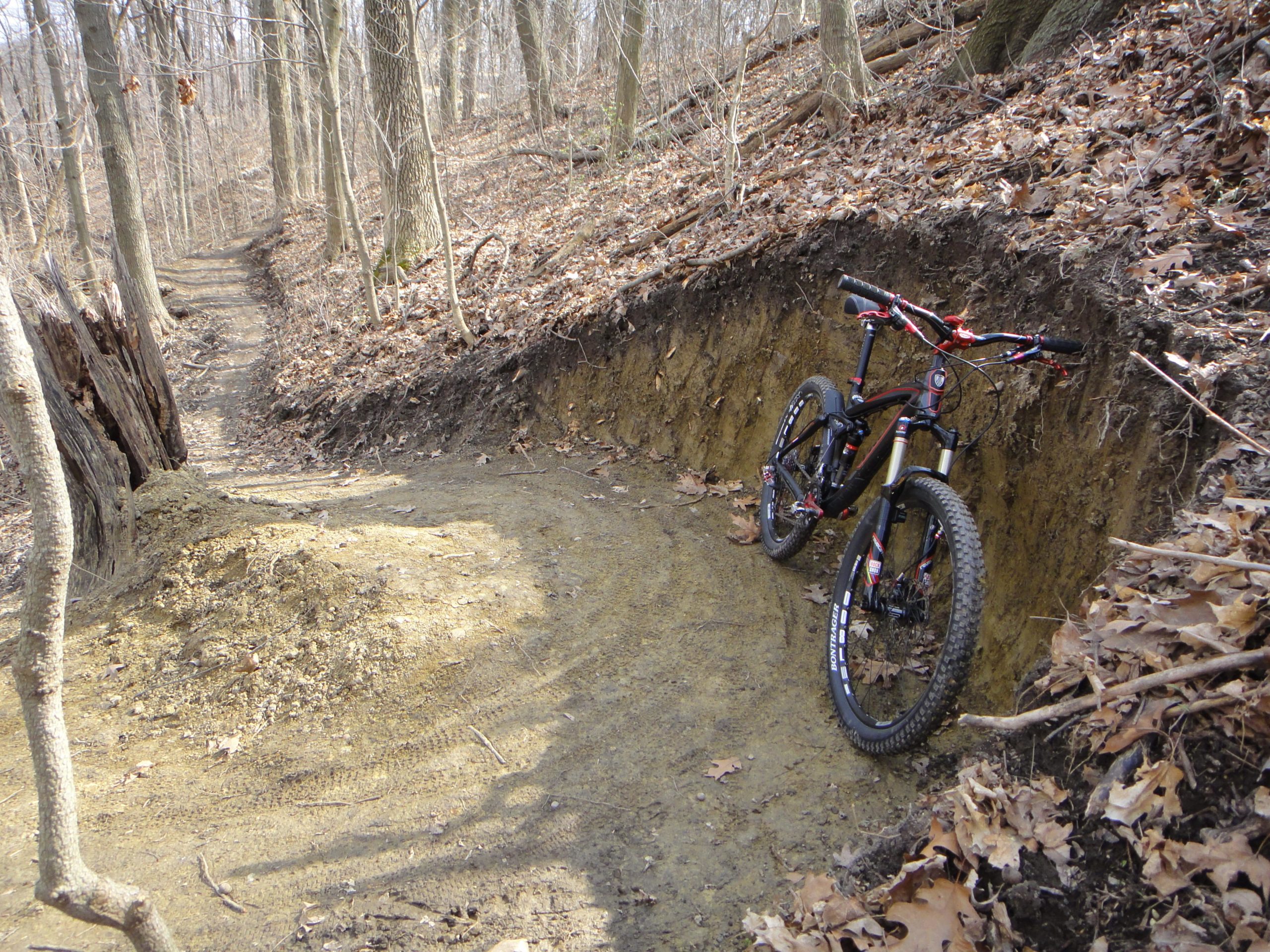 A mountain bike resting against a dirt wall on a forest trail, surrounded by dry leaves and bare trees. A dirt pile is visible on the path, indicating recent trail work. The scene shows a winding trail leading deeper into the woods. Kickapoo mountain bike trail.
