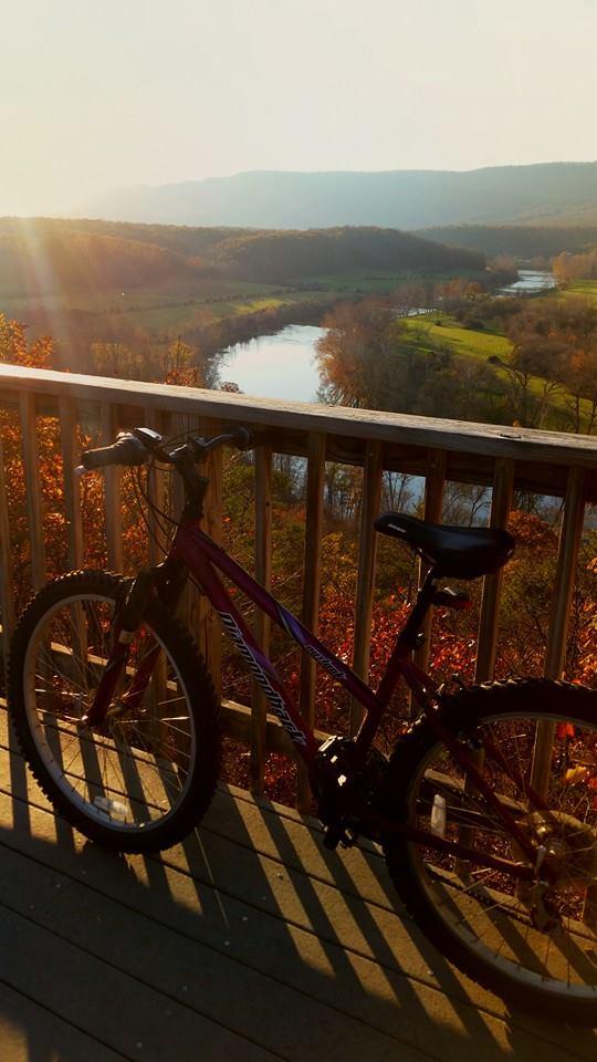 Diamondback Outlook: A mountain bike leaning against a wooden railing, with a scenic view of a river and rolling hills in the background. The scene is bathed in warm sunlight during sunset, highlighting autumn foliage in shades of red and orange.