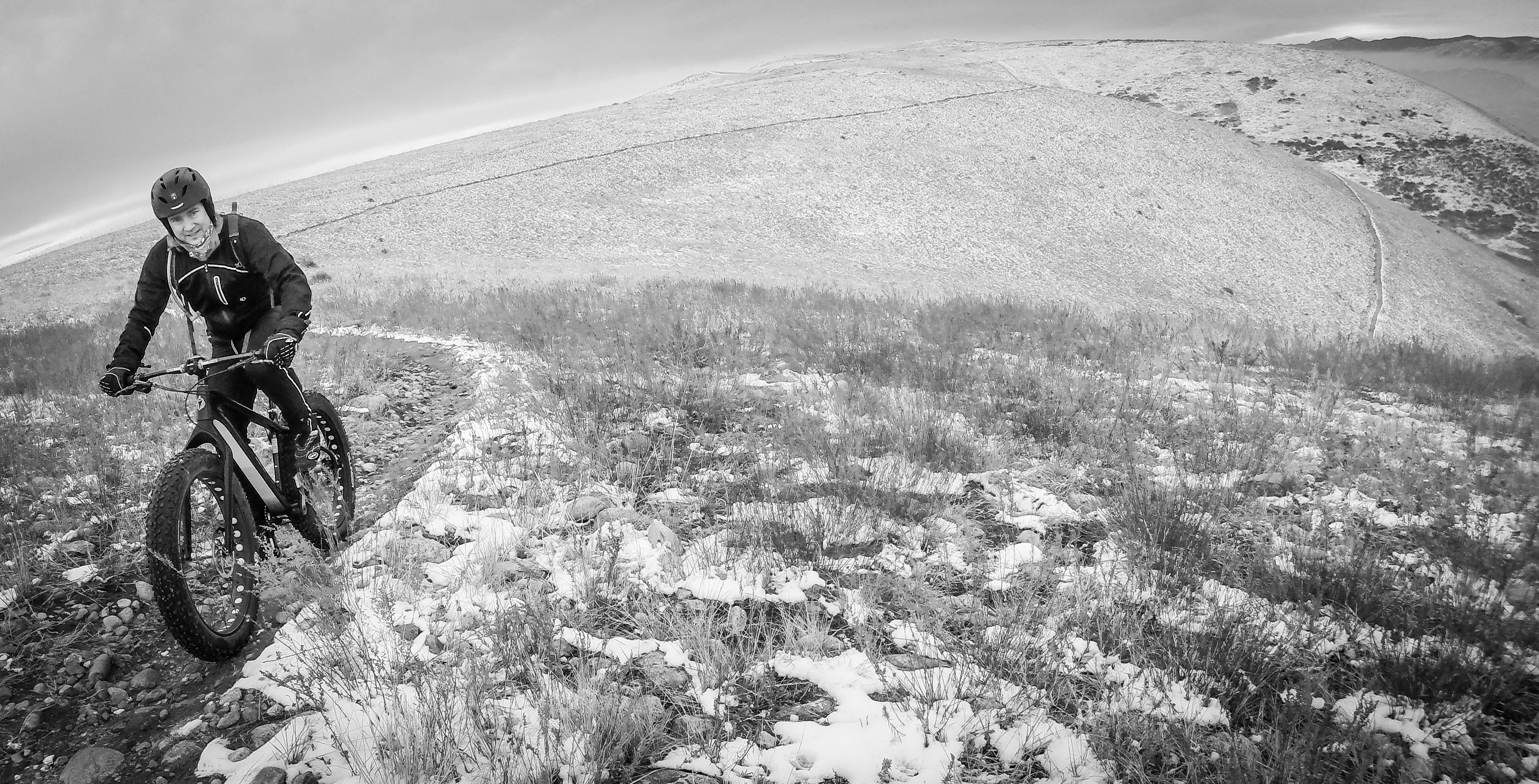 A cyclist riding a fat bike on a snowy, rugged trail, surrounded by grassy terrain and rolling hills, captured in black and white.
