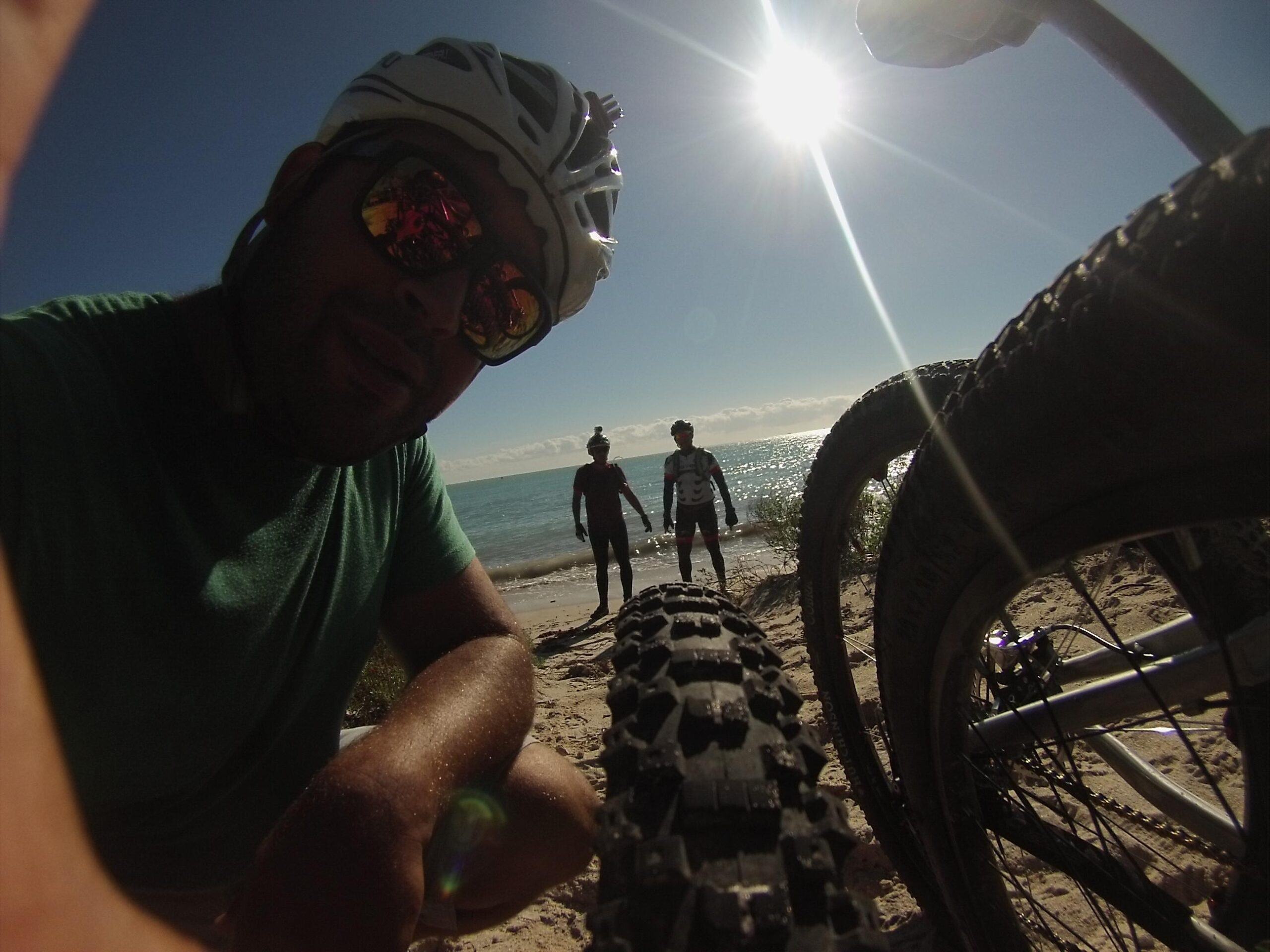 A close-up selfie of a cyclist wearing a helmet and sunglasses, with beach scenery in the background. Two other cyclists are seen standing near the shore, with bicycles partially visible in the foreground, emphasizing the outdoor adventure vibe. The sun shines brightly in the sky over the ocean. Virginia Key North Point mountain bike trail.