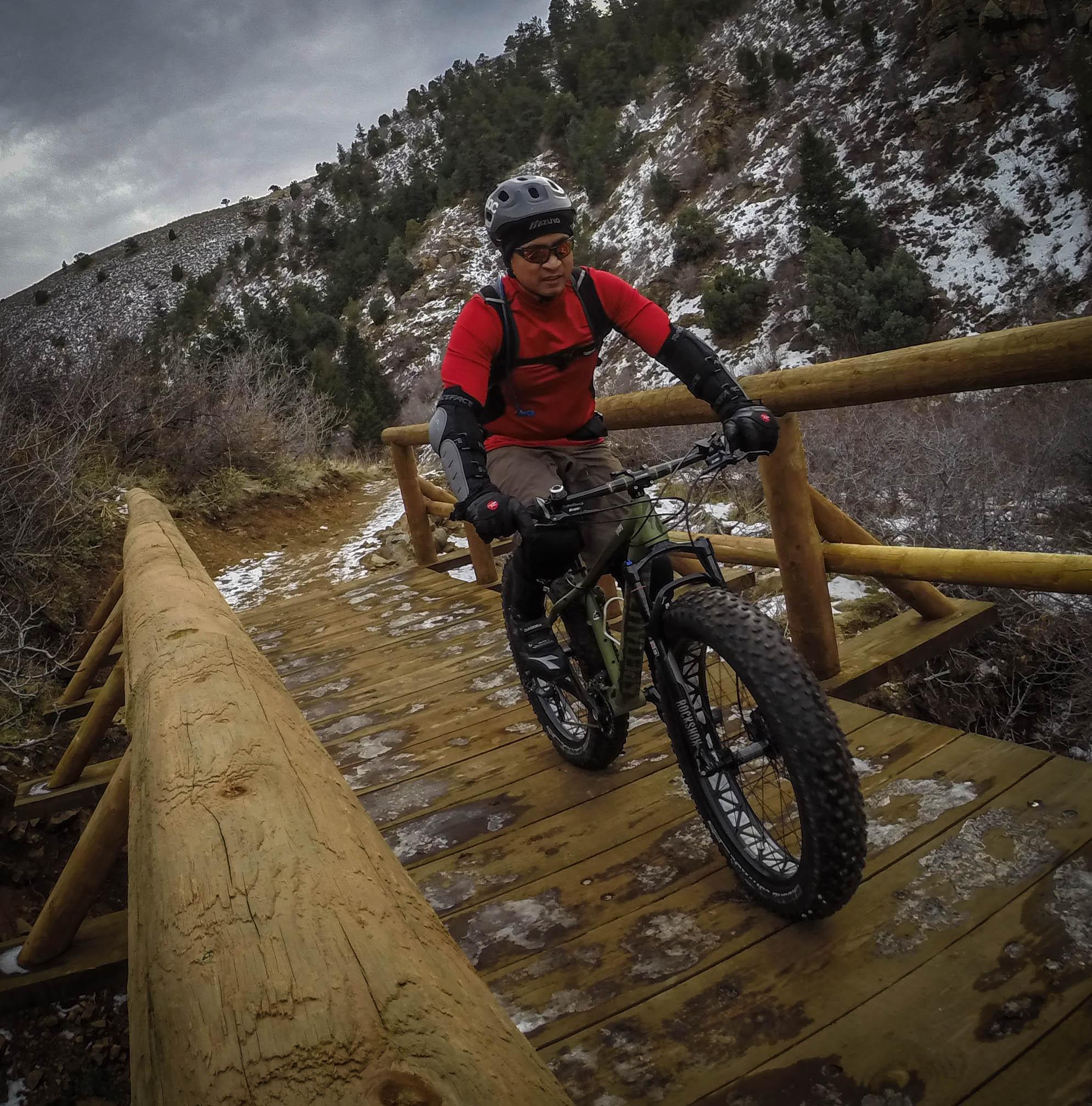 A cyclist wearing a helmet and protective gear rides a mountain bike over a wooden bridge in a natural landscape. The scene features a backdrop of hills with snow patches and sparse trees, under a cloudy sky. Apex Park mountain bike trail.