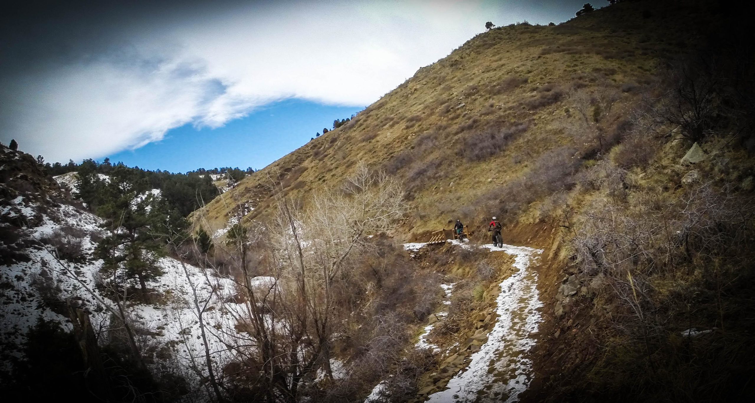 Two cyclists riding on a narrow trail in a mountainous area. The landscape features a mix of snow and dry terrain, with rocky slopes and patches of green vegetation. Above, a blue sky with scattered clouds enhances the natural beauty of the scene. Apex Park mountain bike trail.