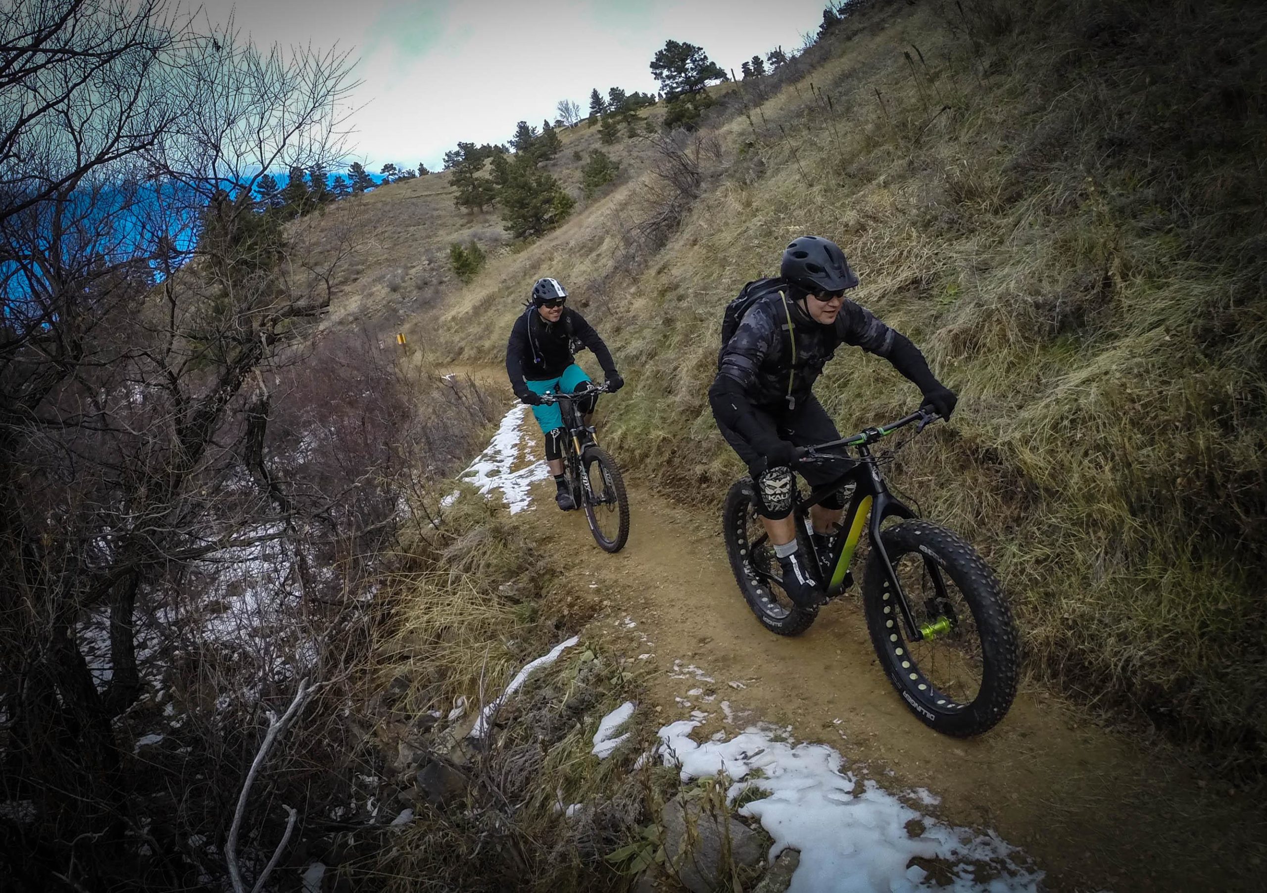 Two mountain bikers traversing a dirt trail lined with sparse vegetation and patches of snow. The landscape features rolling hills and a blue sky with scattered clouds. One rider is wearing a black jacket and helmet, while the other is in a black sweatshirt and turquoise shorts. Both are focused on navigating the trail. Apex Park mountain bike trail.