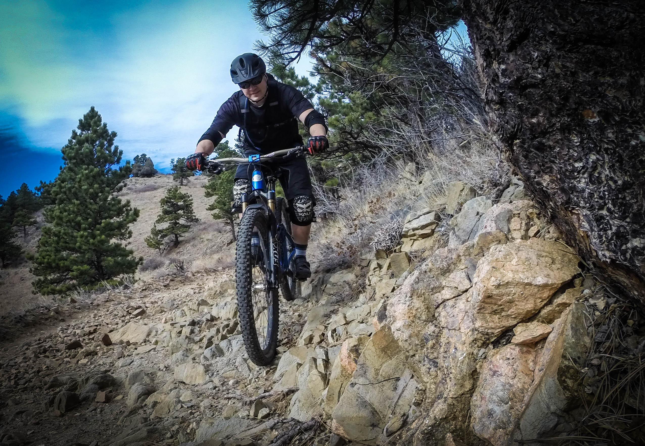 A mountain biker riding on a rocky trail surrounded by trees, with a mix of blue sky and clouds in the background. The rider is focused, wearing a helmet and protective gear, as they navigate a challenging terrain. White Ranch mountain bike trail.