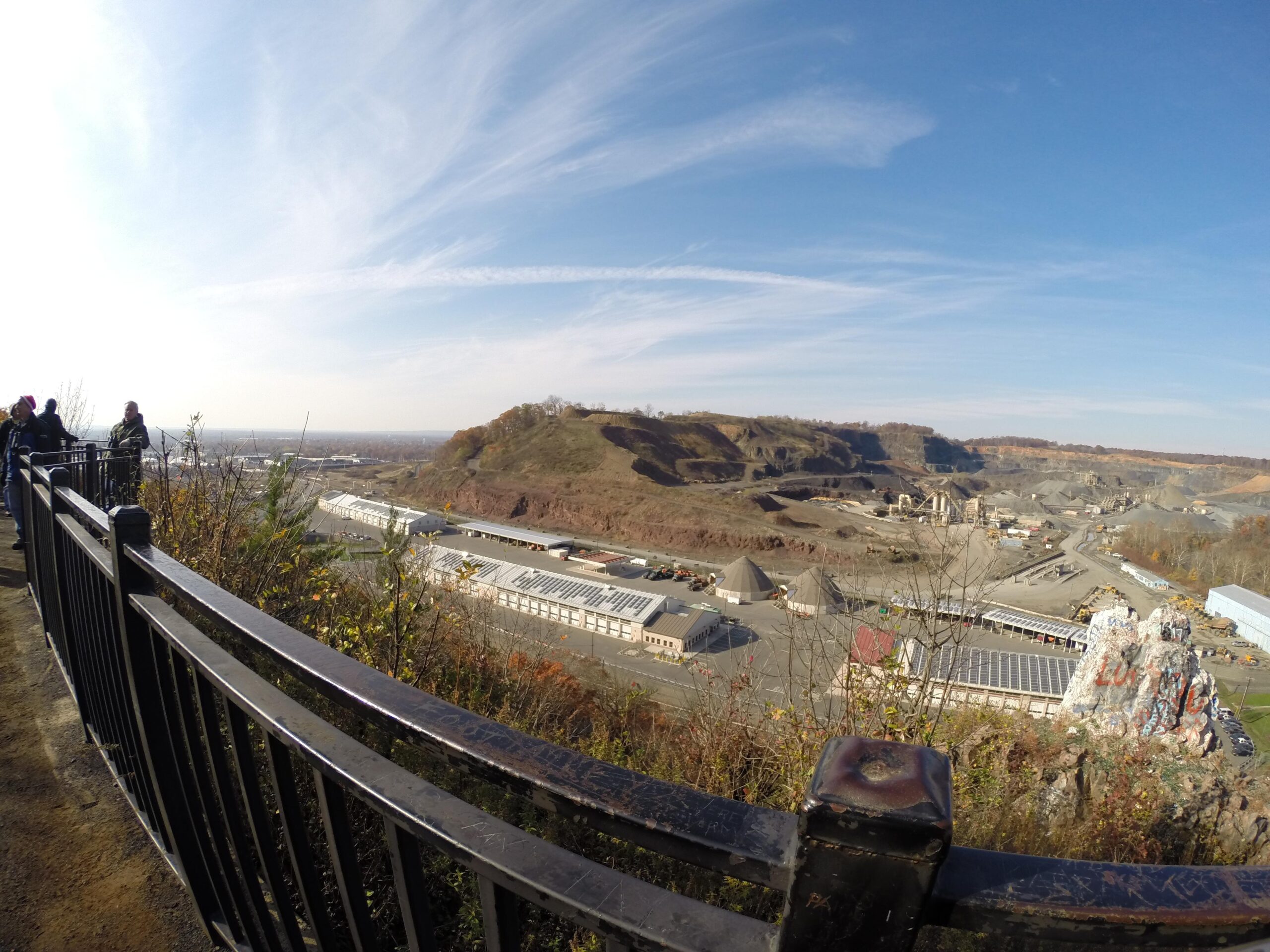 A panoramic view of a mining site taken from a viewing area. In the foreground, a black railing and some vegetation are visible, while several people can be seen standing by the railing, observing the landscape. The background features a large quarry with various machinery and piles of materials. The sky is clear with scattered clouds, showcasing a blue atmosphere. Chimney Rock mountain bike trail.