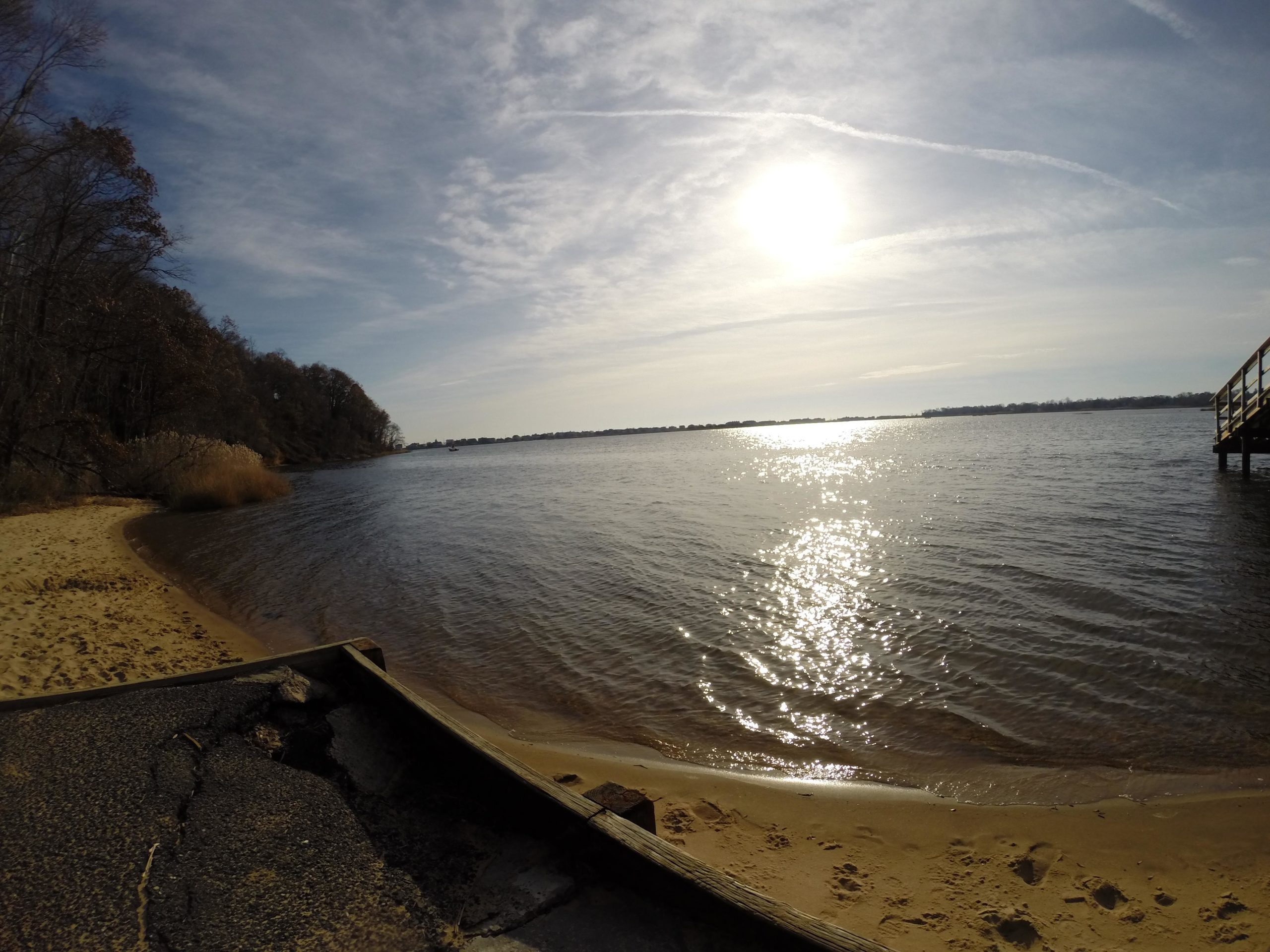 A scenic view of a calm river or lake with a sandy shore, surrounded by trees. The sun is shining brightly in the sky, creating reflections on the water's surface. A wooden pier or dock is visible on the right side of the image, adding to the tranquil atmosphere. Hartshorne Woods Park mountain bike trail.