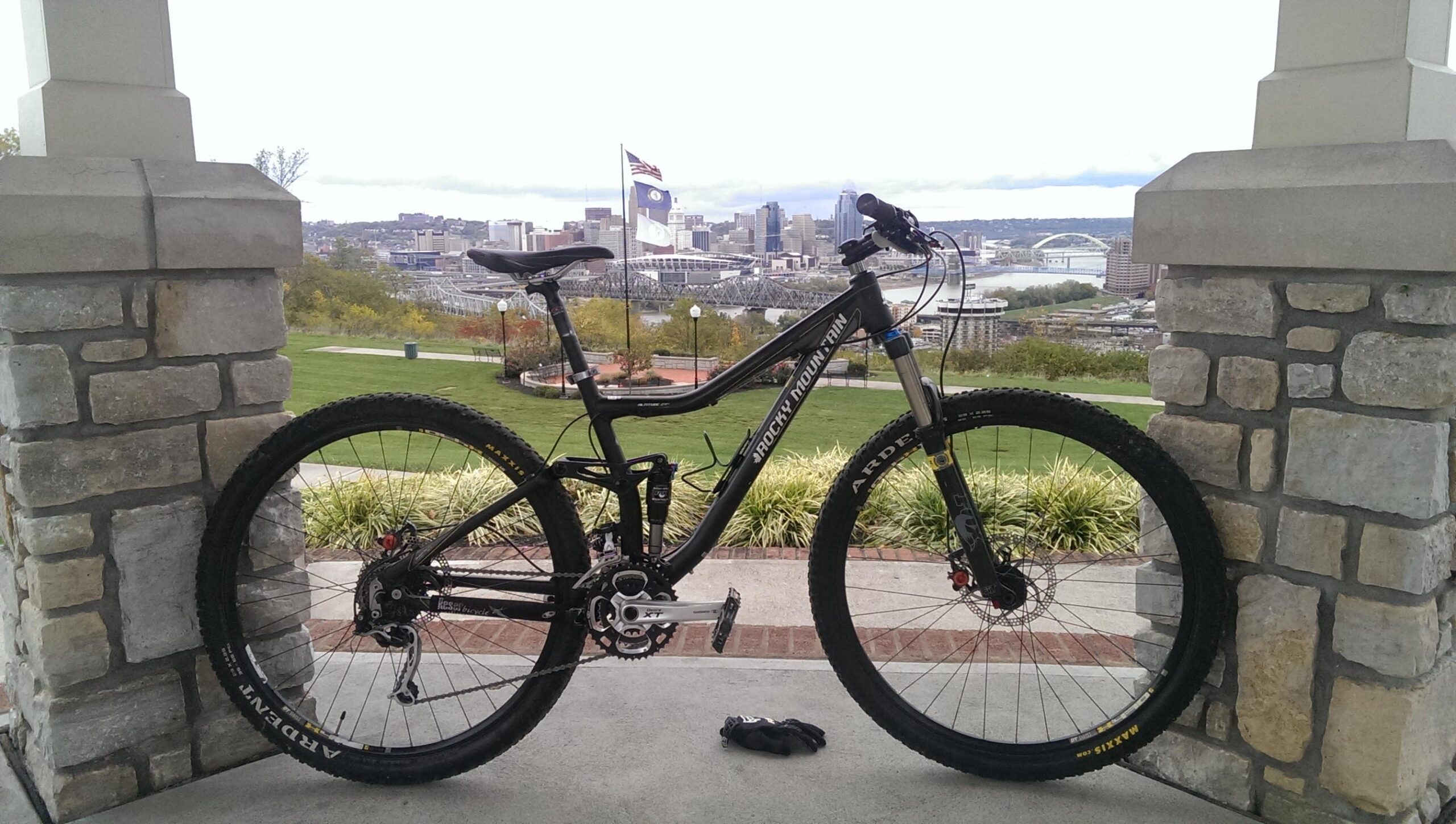 Rocky Mountain Altitude 29se: A mountain bike leaning against a stone pillar with a city skyline in the background. The scene captures a green park area with trees and a few scattered buildings. A cloudy sky is visible above, and an American flag can be seen in the distance.