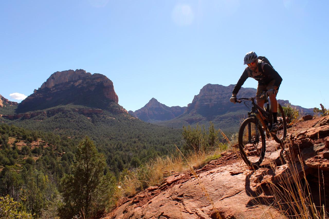 A mountain biker navigating a rocky trail with a scenic backdrop of red rock formations and a clear blue sky. Vegetation surrounds the trail, adding to the natural landscape. Upper Dry Creek Area Trails mountain bike trail.