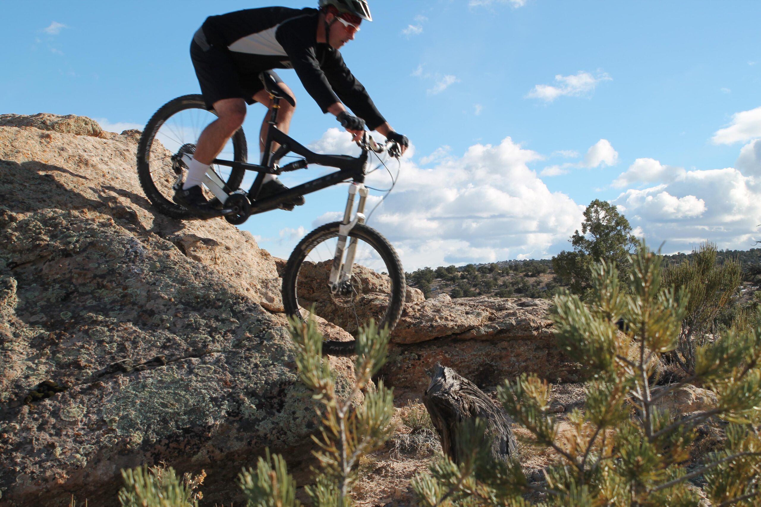 A mountain biker navigating a rocky terrain, airborne over a large boulder, with a blue sky and clouds in the background. Lunch Line mountain bike trail.