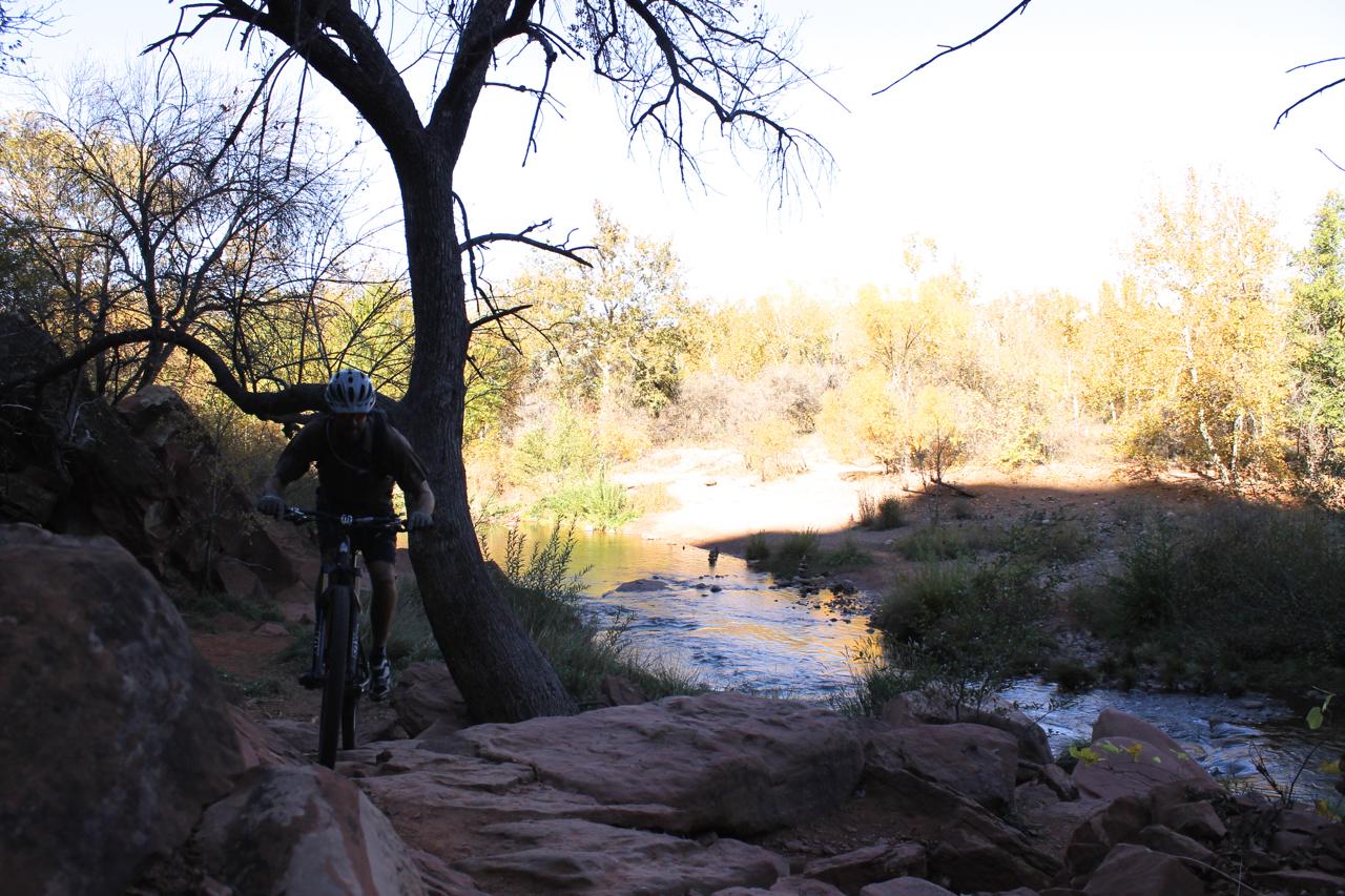A mountain biker navigates a rocky trail next to a winding creek, surrounded by autumn foliage and bare trees. The scene captures the essence of outdoor adventure in a natural setting. Easy Breezy mountain bike trail.