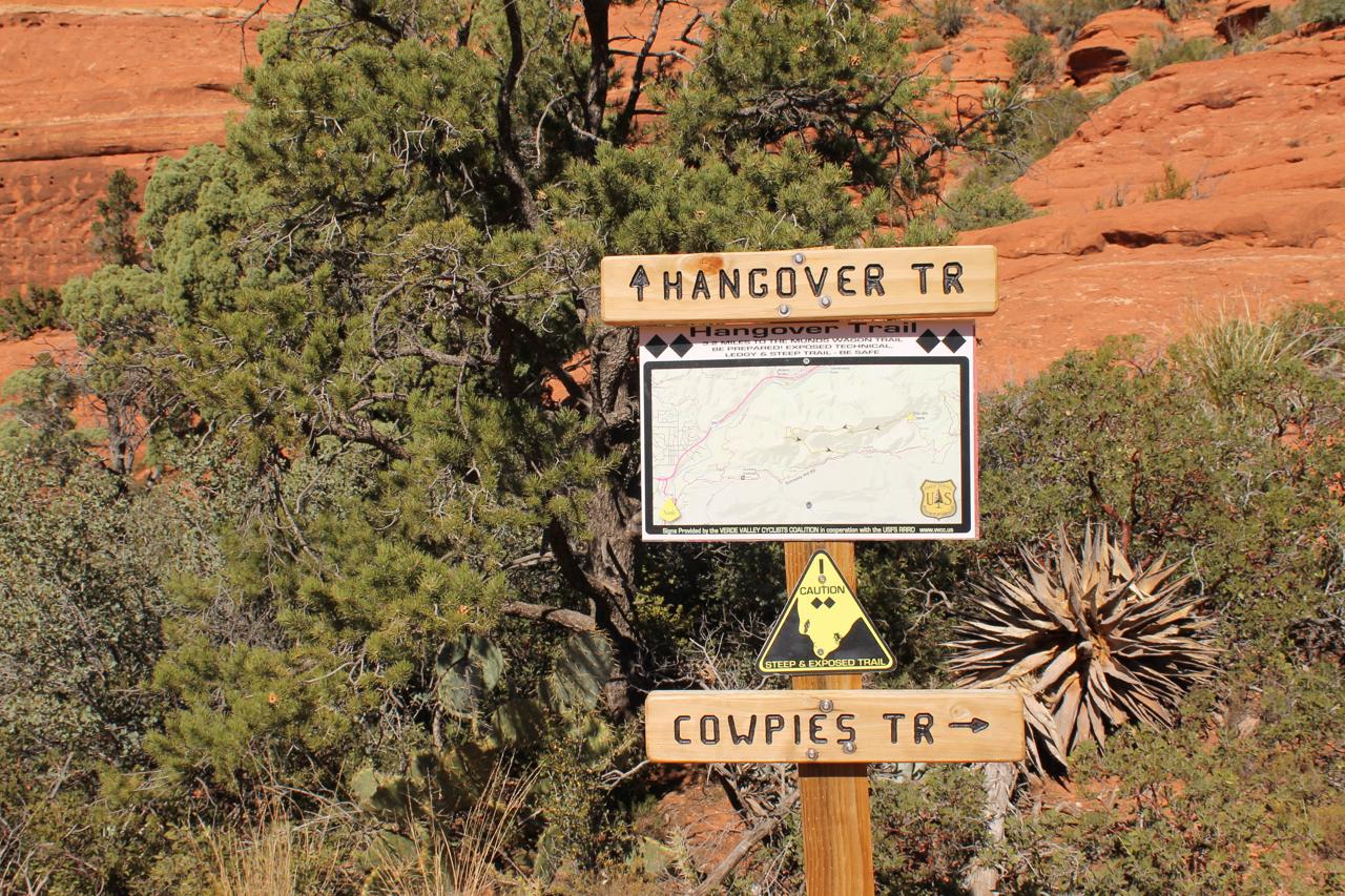 Signpost at a trail intersection in a desert landscape, featuring directions for "Hangover Trail" and "Cowpies Trail." The sign includes a map of the area, with caution symbols indicating steep and exposed terrain. Surrounding vegetation includes trees and shrubs typical of arid environments. Red rock formations are visible in the background. Hangover mountain bike trail.