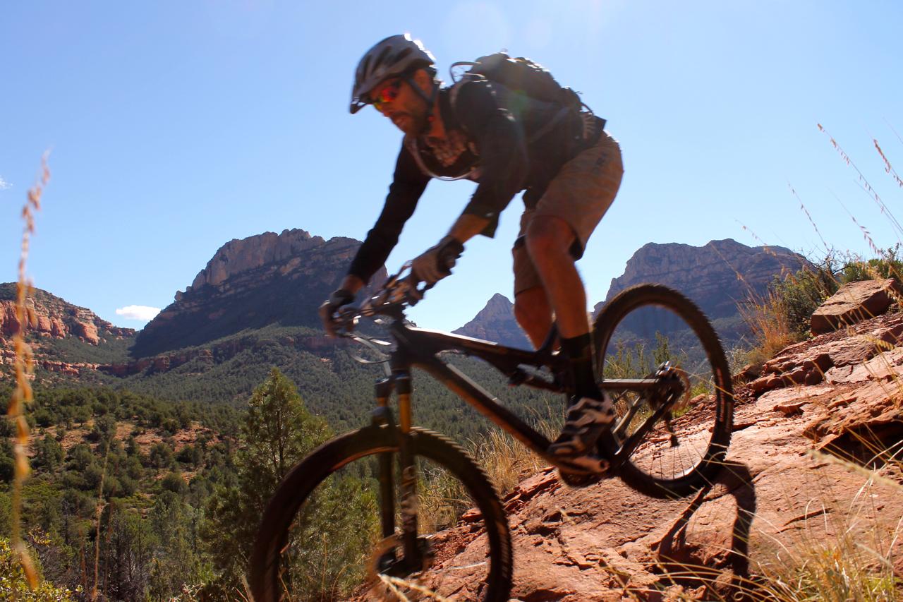 A mountain biker navigating a rocky trail with mountains in the background on a sunny day. The biker is wearing a helmet, sunglasses, and outdoor gear, showcasing an active and adventurous spirit. Upper Dry Creek Area Trails mountain bike trail.