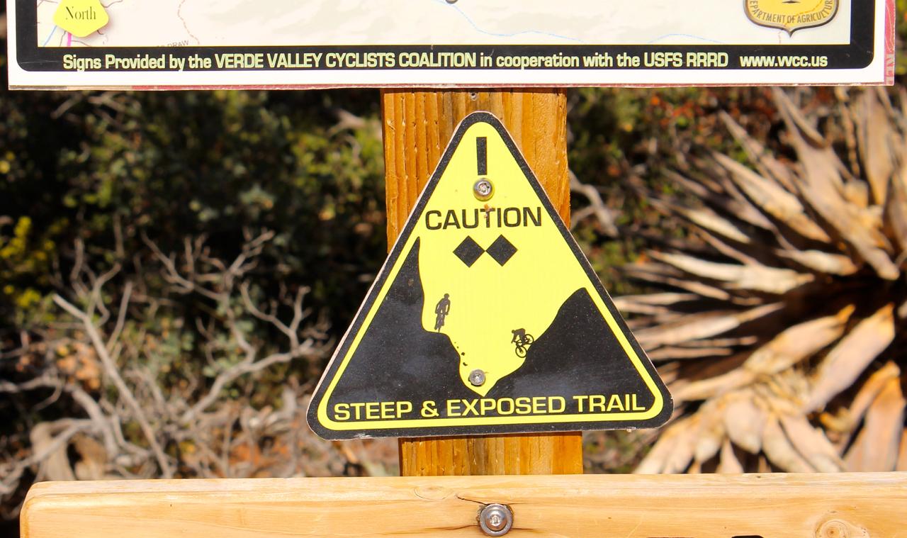 Caution sign indicating a steep and exposed trail, featuring icons of a mountain biker and a steep slope. The sign is affixed to a wooden post, with a background of natural vegetation. Hangover mountain bike trail.