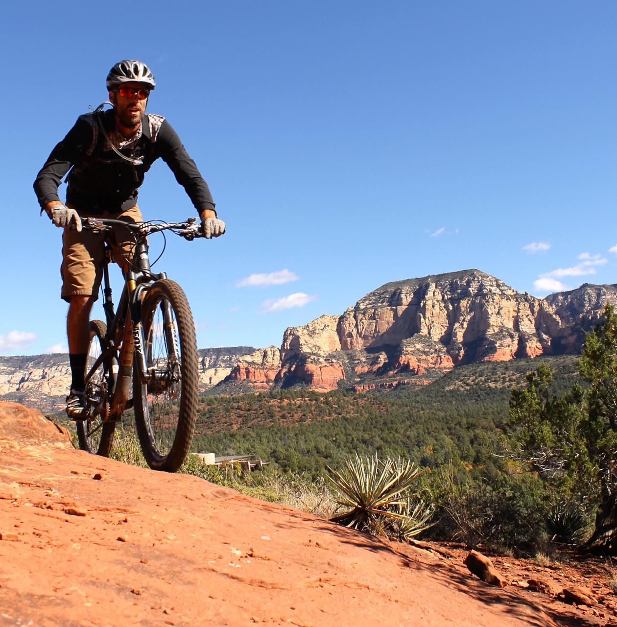 A mountain biker navigating rocky terrain in a desert landscape, with red rock formations and green vegetation in the background under a clear blue sky. Upper Dry Creek Area Trails mountain bike trail.