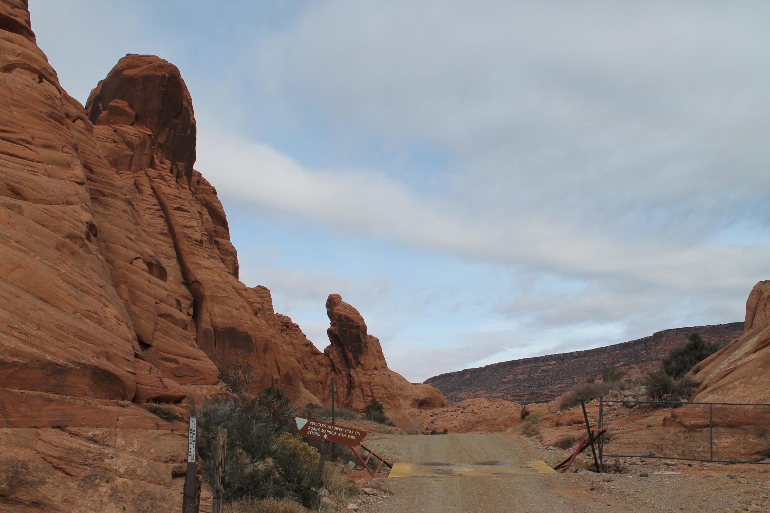 Desert landscape featuring tall, reddish rock formations lining a dirt road, with a cloudy sky in the background. A sign indicating vehicle access is partially visible along the side of the road. Flat Pass mountain bike trail.