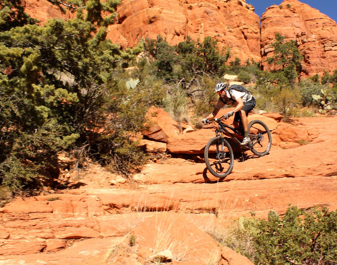 A mountain biker rides over rocky terrain in a desert landscape, surrounded by red sandstone formations and sparse vegetation under a clear blue sky. Hangover mountain bike trail.