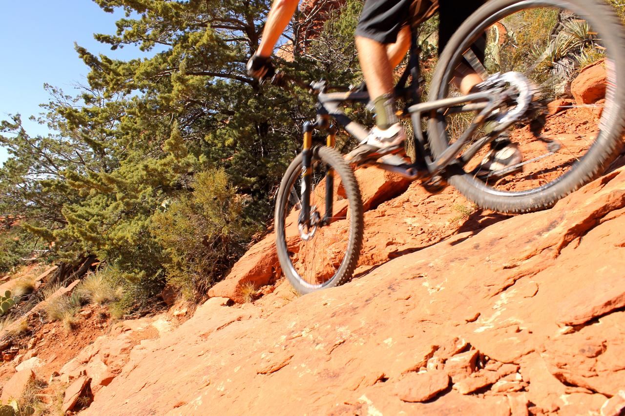 A cyclist maneuvering a mountain bike on a rocky trail, surrounded by greenery and red rock terrain under a clear blue sky. Hangover mountain bike trail.