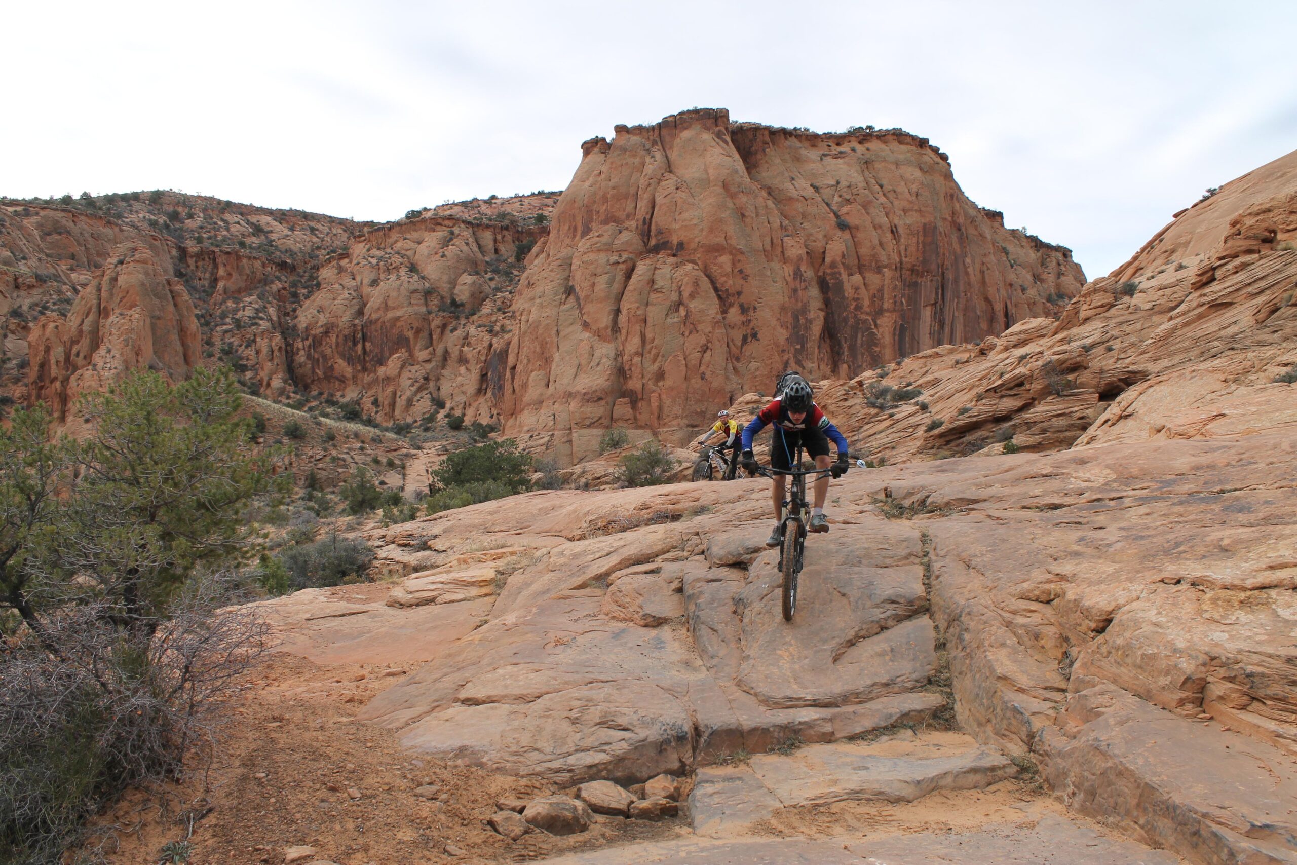 A mountain biker navigating a rocky trail in a scenic desert landscape, featuring reddish rock formations and sparse vegetation in the background. The biker is wearing a helmet and protective gear, showcasing an action shot as they maneuver down the terrain. Flat Pass mountain bike trail.