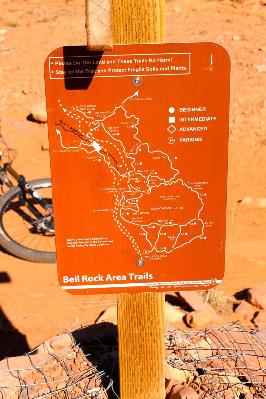 Trail sign for Bell Rock Area, featuring a map of the hiking and biking trails, with markers indicating beginner, intermediate, and advanced routes. The sign advises trail users to protect the land and stay on designated paths. A bike is partially visible in the background, set against a red dirt landscape. Bell Rock Area Trails mountain bike trail.