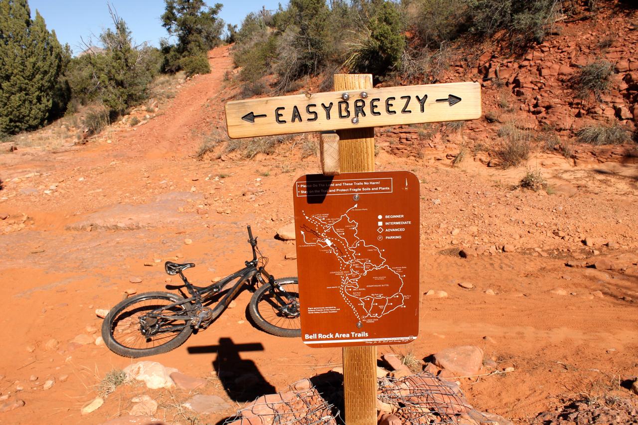 A wooden signpost labeled "EASYBREEZY" points towards a dirt trail surrounded by vegetation. In the foreground, a black mountain bike leans against the sign. A brown informational map is attached to the sign, indicating various trail routes and difficulty levels in the Bell Rock area. The background features a reddish landscape typical of outdoor biking trails. Bell Rock Area Trails mountain bike trail.