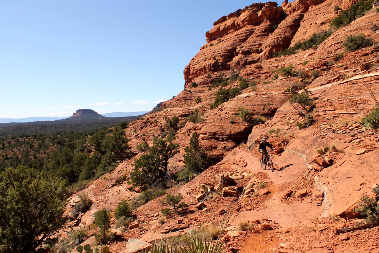 A cyclist navigating a narrow trail along a rocky, red landscape with scattered vegetation and distant mountains under a clear blue sky. Upper Dry Creek Area Trails mountain bike trail.