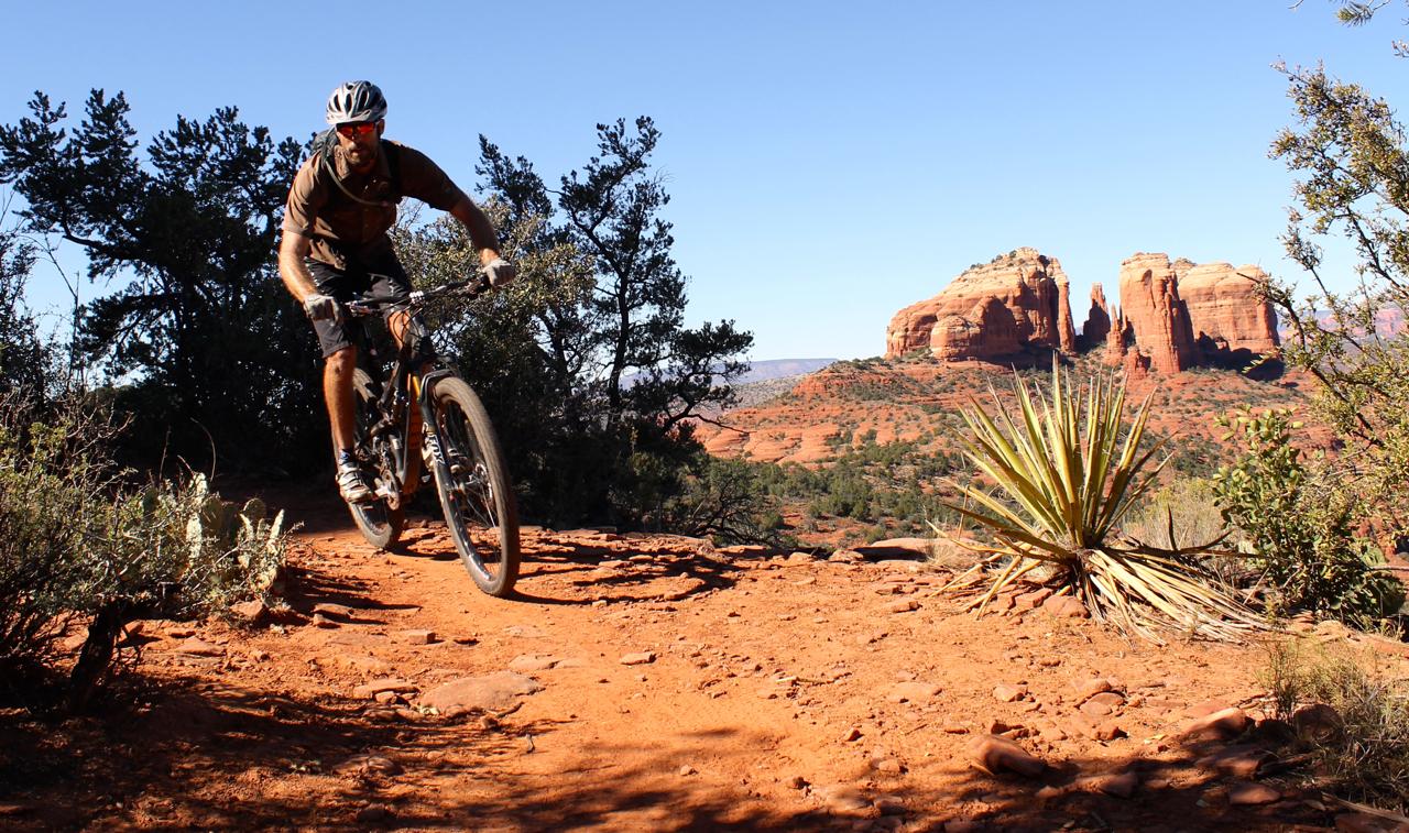 A mountain biker navigates a rocky dirt trail surrounded by desert vegetation, with red rock formations in the background under a clear blue sky. Hiline mountain bike trail.