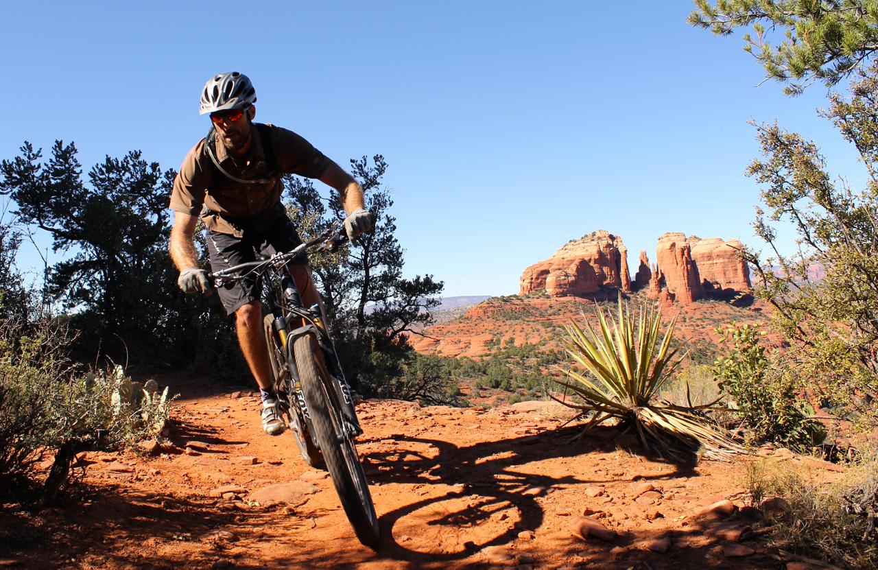 A mountain biker navigating a dirt trail in a desert landscape with red rock formations in the background under a clear blue sky. Hiline mountain bike trail.