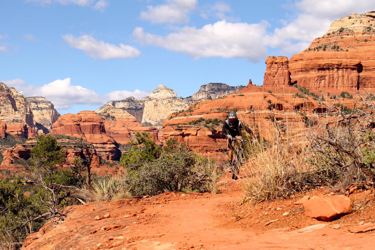 A mountain biker riding along a rocky trail surrounded by red rock formations and greenery, with a clear blue sky and clouds in the background. Upper Dry Creek Area Trails mountain bike trail.