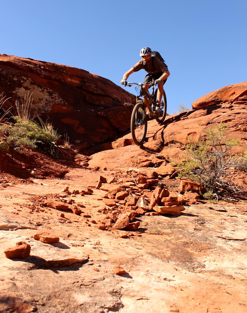 A mountain biker skillfully navigates a rocky terrain under a clear blue sky, leaping off a ledge surrounded by red rock formations and scattered stones. Hiline mountain bike trail.