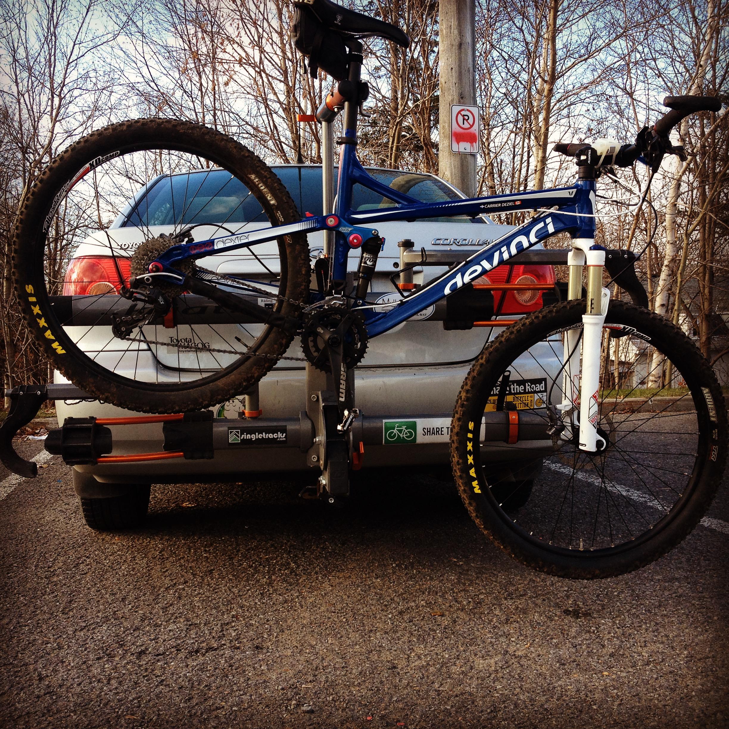 Devinci Troy: A blue mountain bike is mounted on the back of a silver car, with its wheels resting on a bike rack. The bike has visible mud on its tires, indicating recent use. Surrounding the scene are bare trees and a parking sign in the background, suggesting an outdoor location. The car's rear features various stickers, including those promoting cycling.