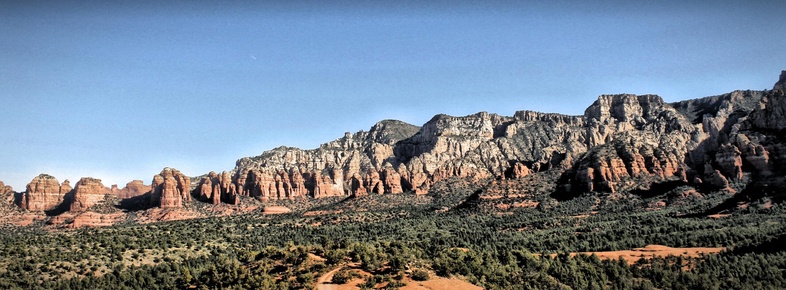 A panoramic view of rugged red rock formations and cliffs under a clear blue sky, surrounded by a landscape of green trees and earthy tones. The scene captures the natural beauty of a desert-like environment, highlighting the unique geological features of the area. Broken Arrow Trail System mountain bike trail.