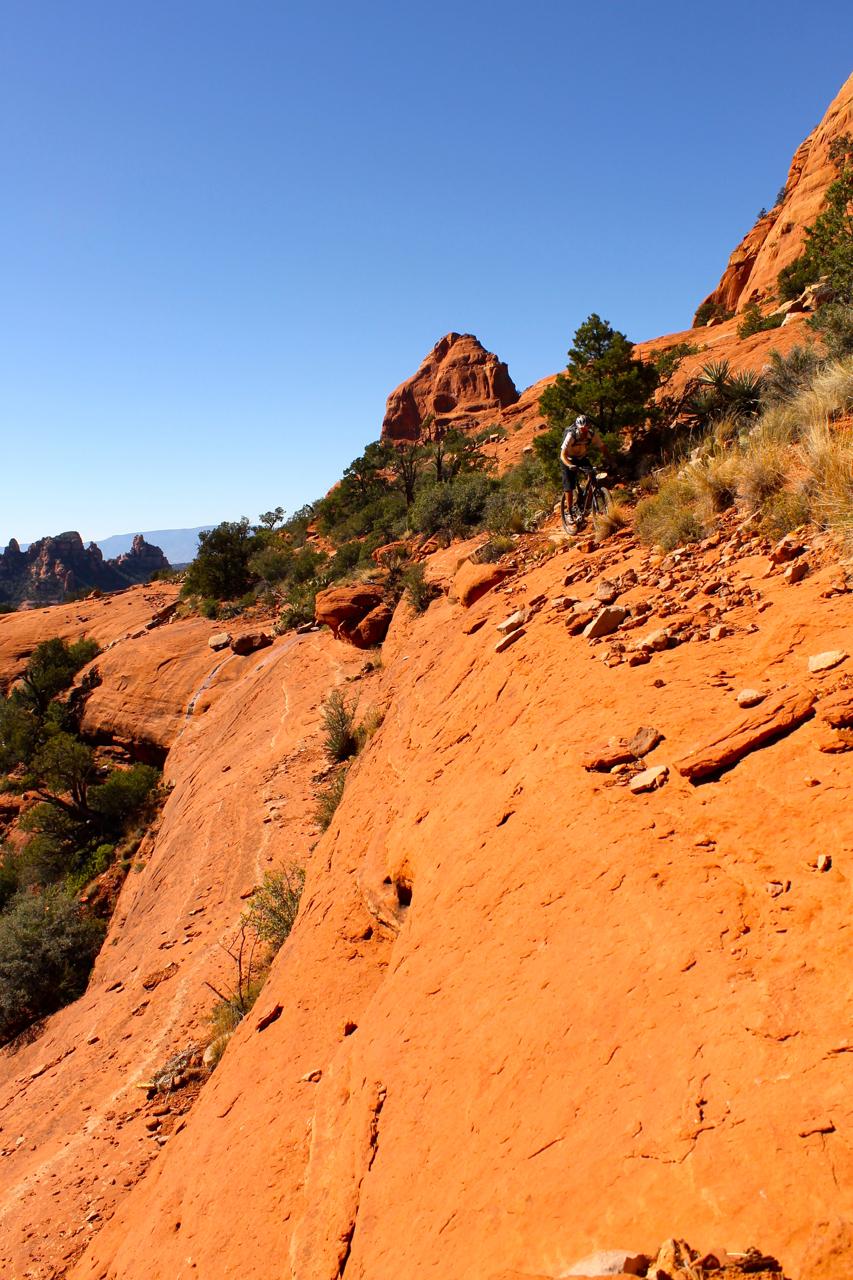 A mountain biker navigating a rocky, orange-hued trail surrounded by sparse vegetation under a clear blue sky. The landscape features distinctive rock formations and a steep incline, highlighting the rugged terrain. Hangover mountain bike trail.