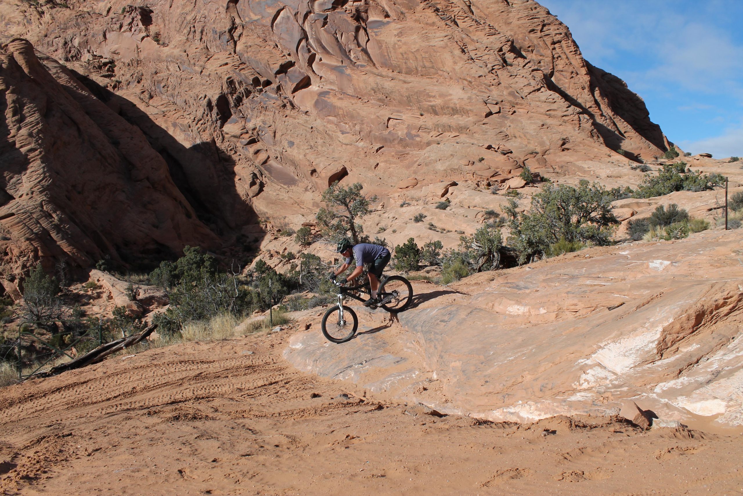 A mountain biker navigating rocky terrain in a desert landscape, with layered reddish-brown rock formations in the background and sparse vegetation. The cyclist is focused and leaning forward on the bike as they descend a slope. Flat Pass mountain bike trail.