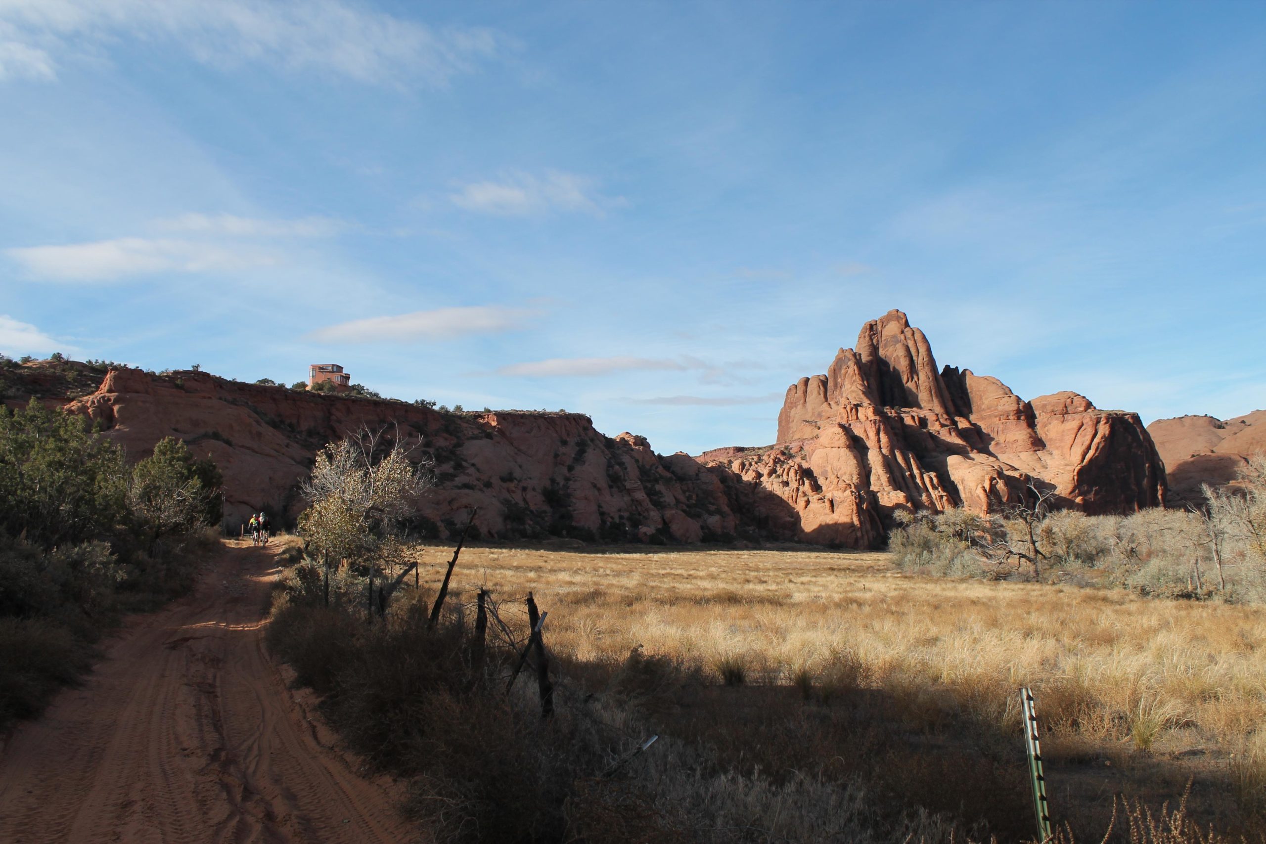 A dirt path leads through a grassy landscape with scrubby vegetation, surrounded by striking red rock formations under a partly cloudy sky. In the distance, two figures can be seen biking along the path, and a rustic structure is perched on a cliff. The scene captures the natural beauty of the area, highlighting the contrasting textures of the terrain. Flat Pass mountain bike trail.