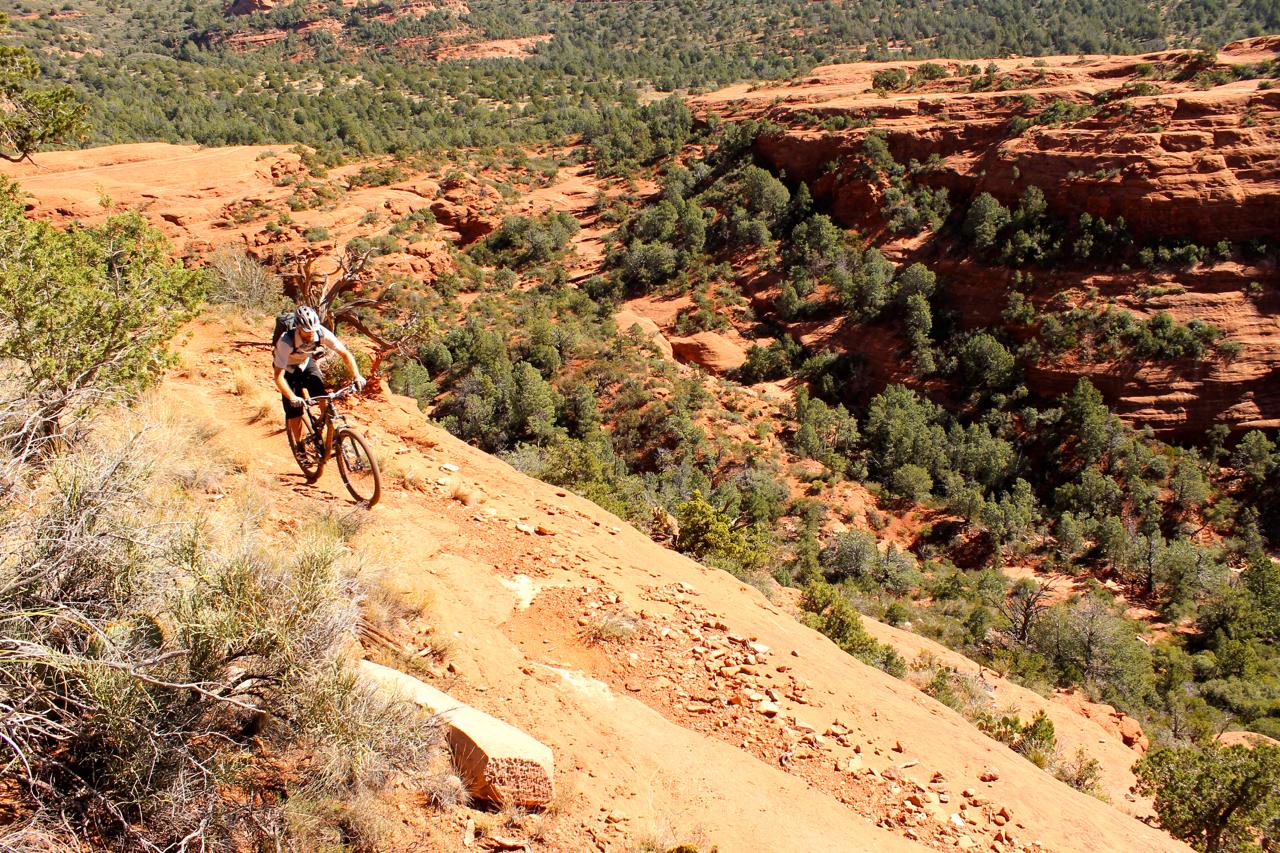 A mountain biker rides down a steep, rocky trail surrounded by red dirt and green vegetation. The landscape features rugged terrain and a scenic view of the valley below. Hangover mountain bike trail.