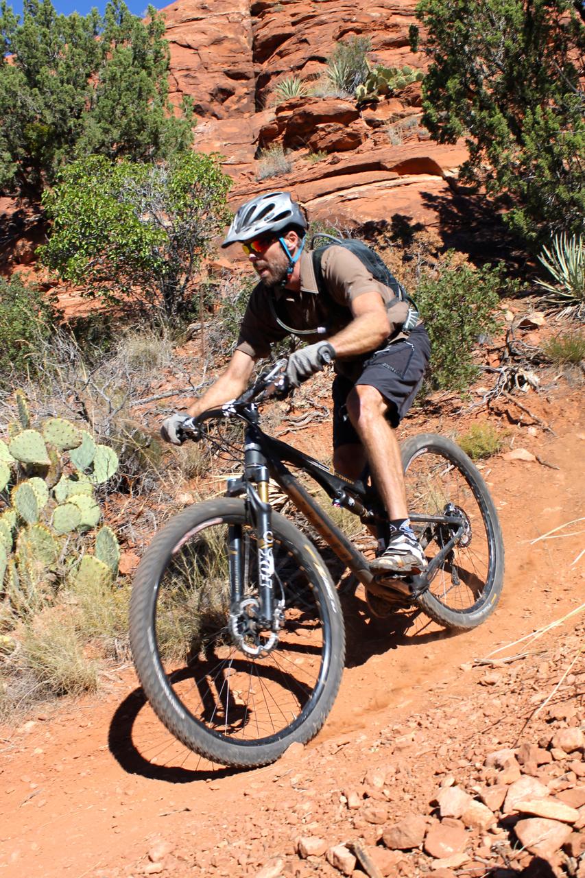 A mountain biker riding on a rocky, dirt trail surrounded by desert vegetation, including cacti and shrubs. The cyclist is wearing a helmet and sunglasses, with a focused expression as he navigates the terrain. Bright blue sky and striking red rock formations are visible in the background. Hiline mountain bike trail.