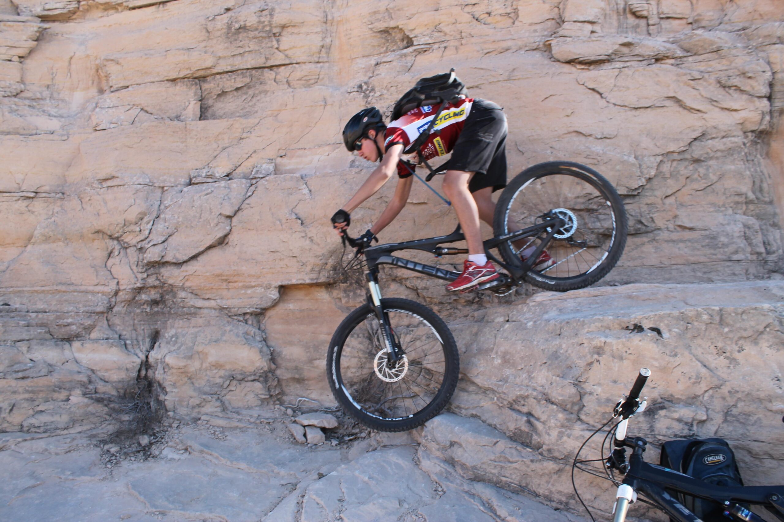 A mountain biker navigating a rocky terrain, balancing on a ledge while descending. The cyclist is wearing a helmet and sporting a jersey, with another bike and a backpack visible nearby on the ground. Mary's Loop / Horsethief Bench mountain bike trail.