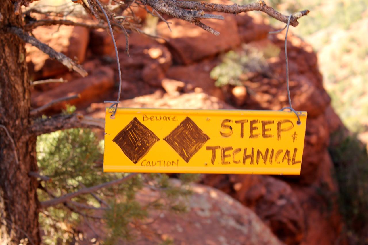 A yellow warning sign hanging from a branch in a rocky outdoor setting, displaying the words "STEAP & TECHNICAL" with additional notes saying "Beware" and "Caution." The background features red rock formations and greenery, indicating a steep and potentially challenging trail. Hiline mountain bike trail.