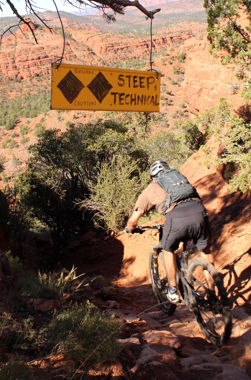 A mountain biker descending a rocky trail near a warning sign that reads "Beware: Steep & Technical." The backdrop features red rock formations and green vegetation, indicating a rugged outdoor environment. Hiline mountain bike trail.