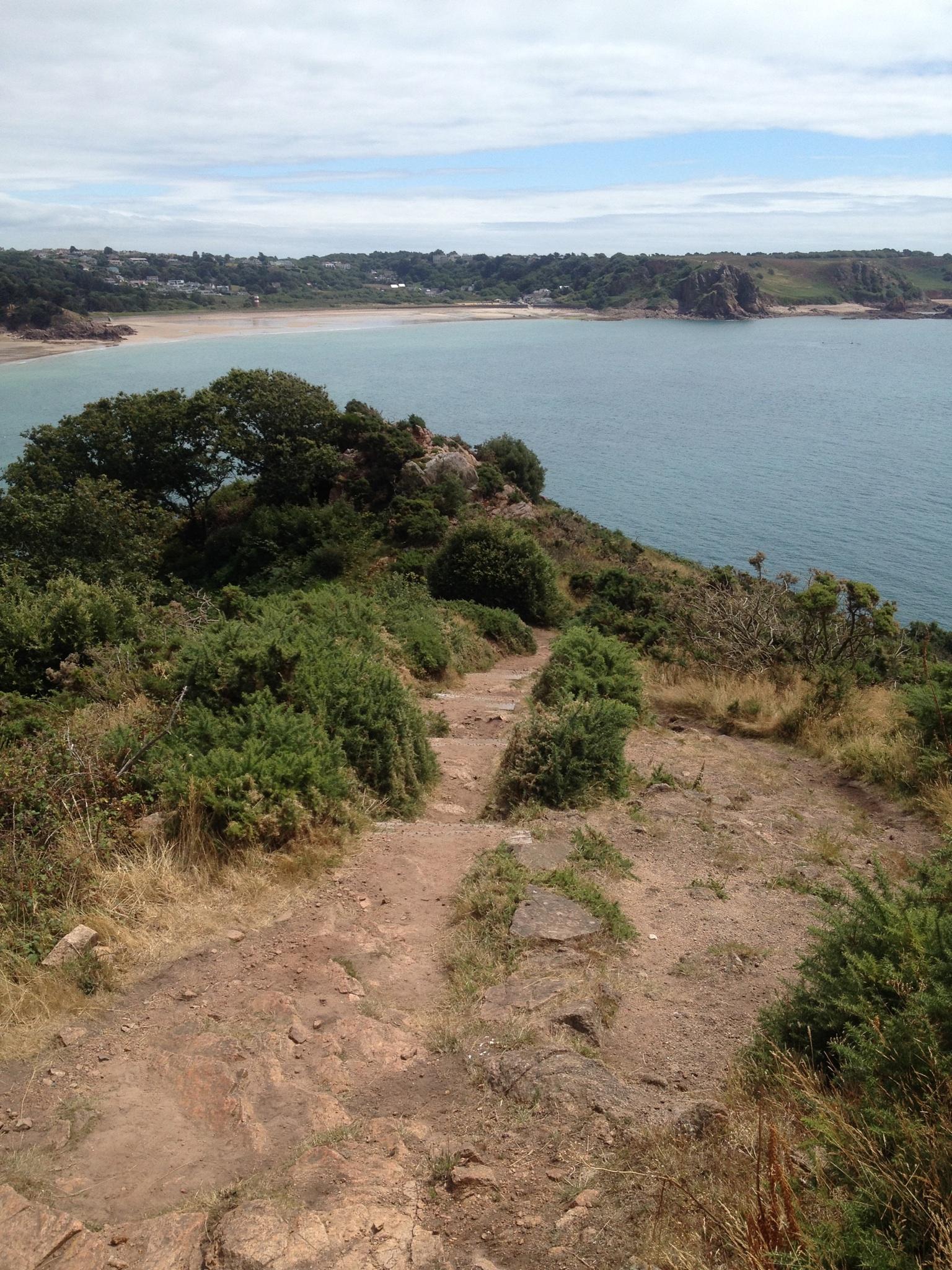 A scenic view from a rocky hillside overlooking a calm bay, with sandy beach areas and lush greenery in the foreground. The coastline features gentle waves and a backdrop of distant hills under a partly cloudy sky. A dirt pathway winds down the hillside, surrounded by shrubs and grass. St Brelade loop smash mountain bike trail.