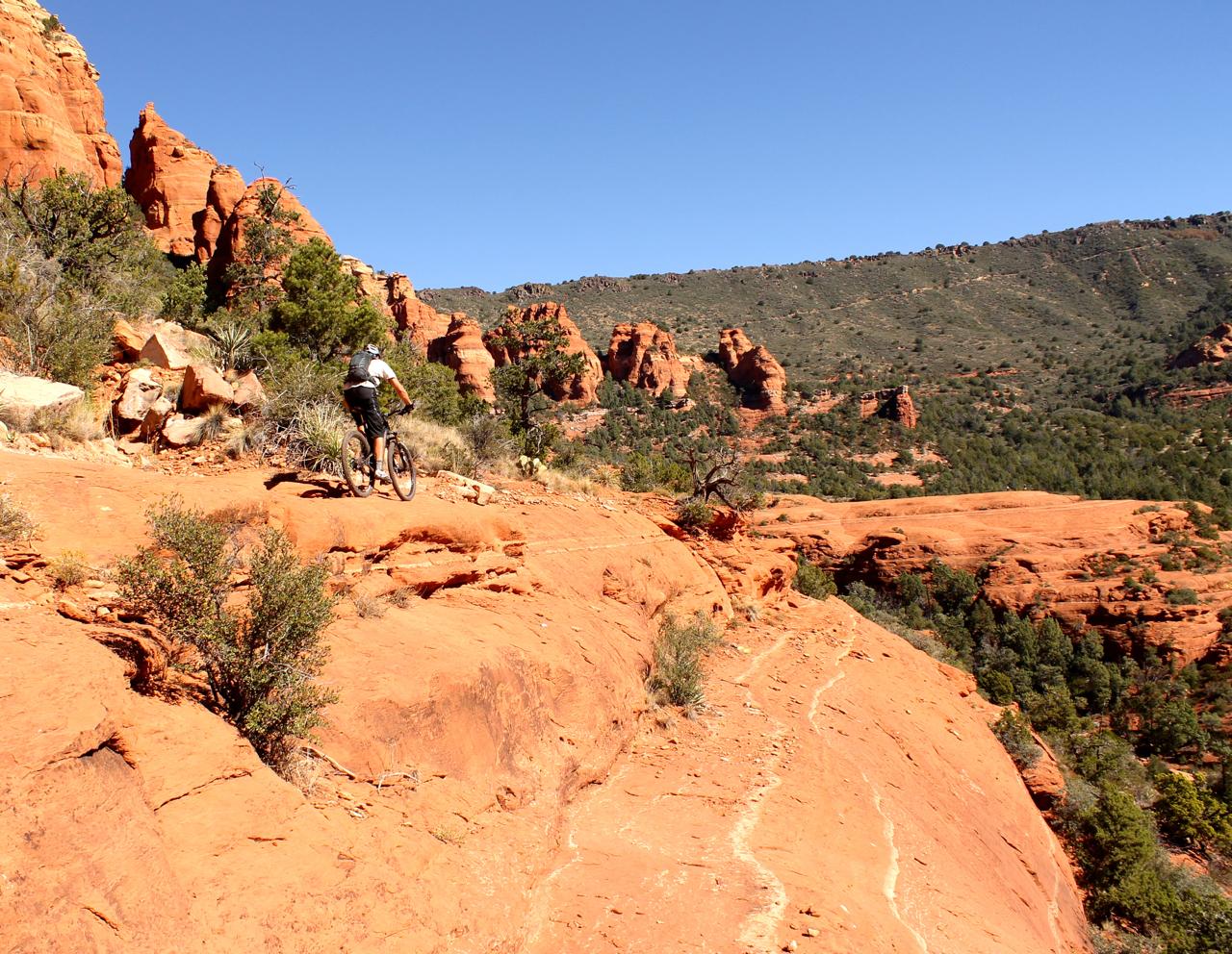 A mountain biker riding along a narrow, rocky trail on a red sandstone landscape, with towering red rock formations and green vegetation in the background under a clear blue sky. Hangover mountain bike trail.