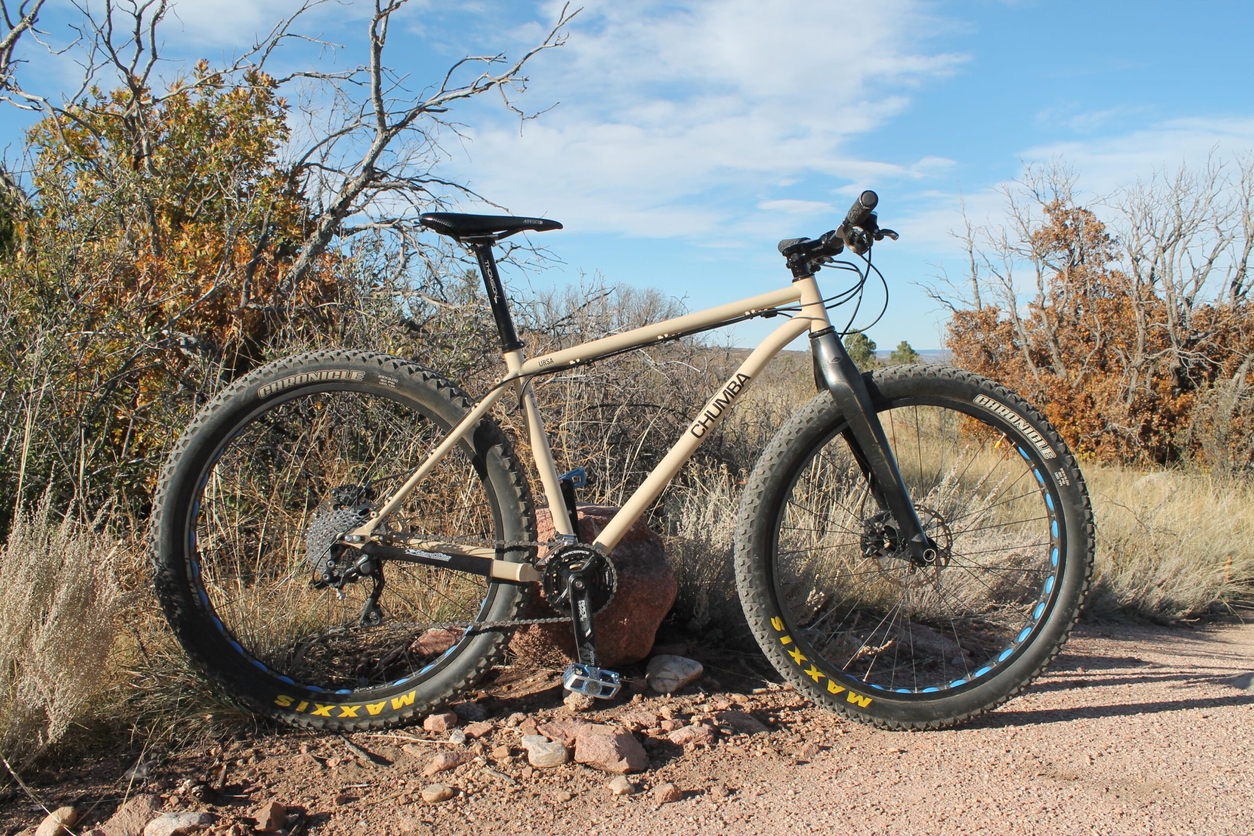 Chumba Ursa: A mountain bike with a tan frame, large knobby tires, and a prominent brand label is positioned on a rocky trail surrounded by dry foliage and sparse trees. The sky is clear with a few clouds, indicating a sunny day in a natural landscape.