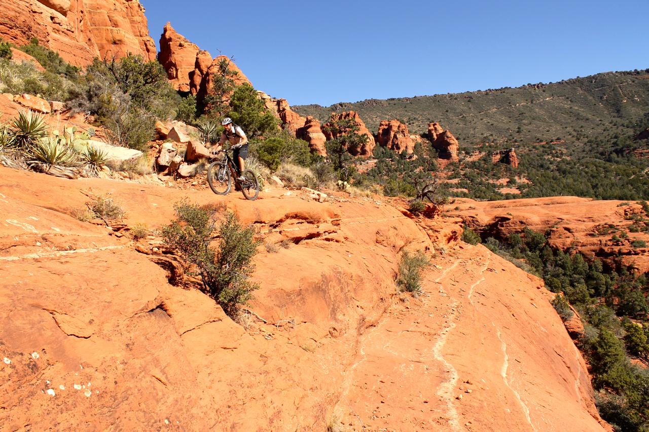 A mountain biker navigating a rocky trail on red sandstone terrain, surrounded by rugged cliffs and greenery under a clear blue sky. Hangover mountain bike trail.
