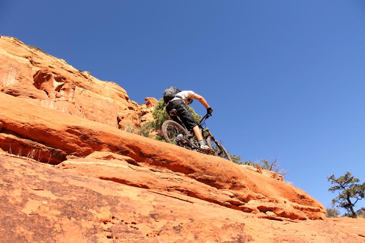 A person riding a mountain bike on a rocky, orange terrain under a clear blue sky. The cyclist is positioned on a slope with rugged rocks and sparse vegetation in the background. Hangover mountain bike trail.